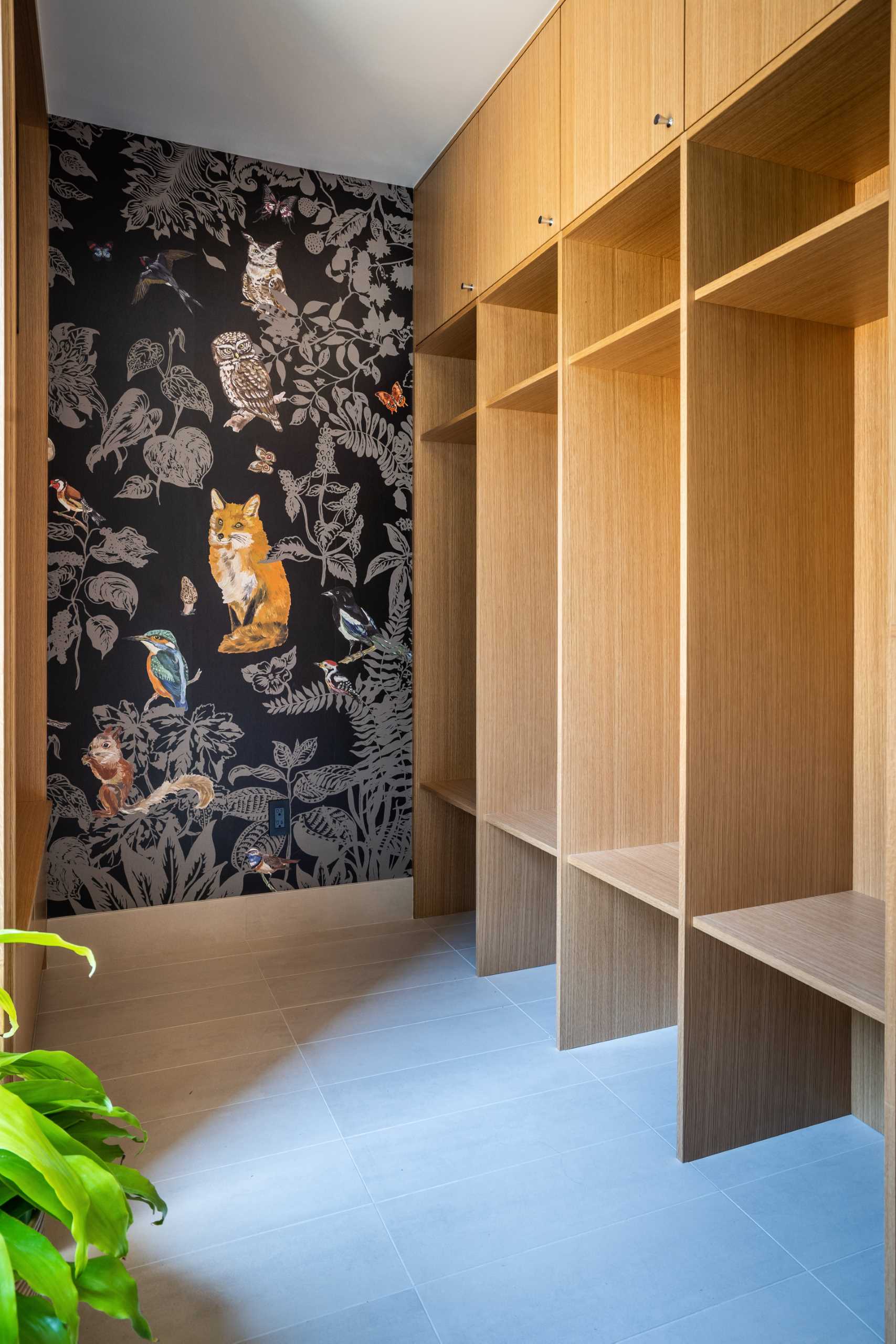 In this mudroom, custom floor-to-ceiling cabinets and shelving take up the wall, while a nature-inspired wallpaper adds an artistic element. Opposite the cabinetry is a pegboard wall that includes ،oks that can be moved when needed.