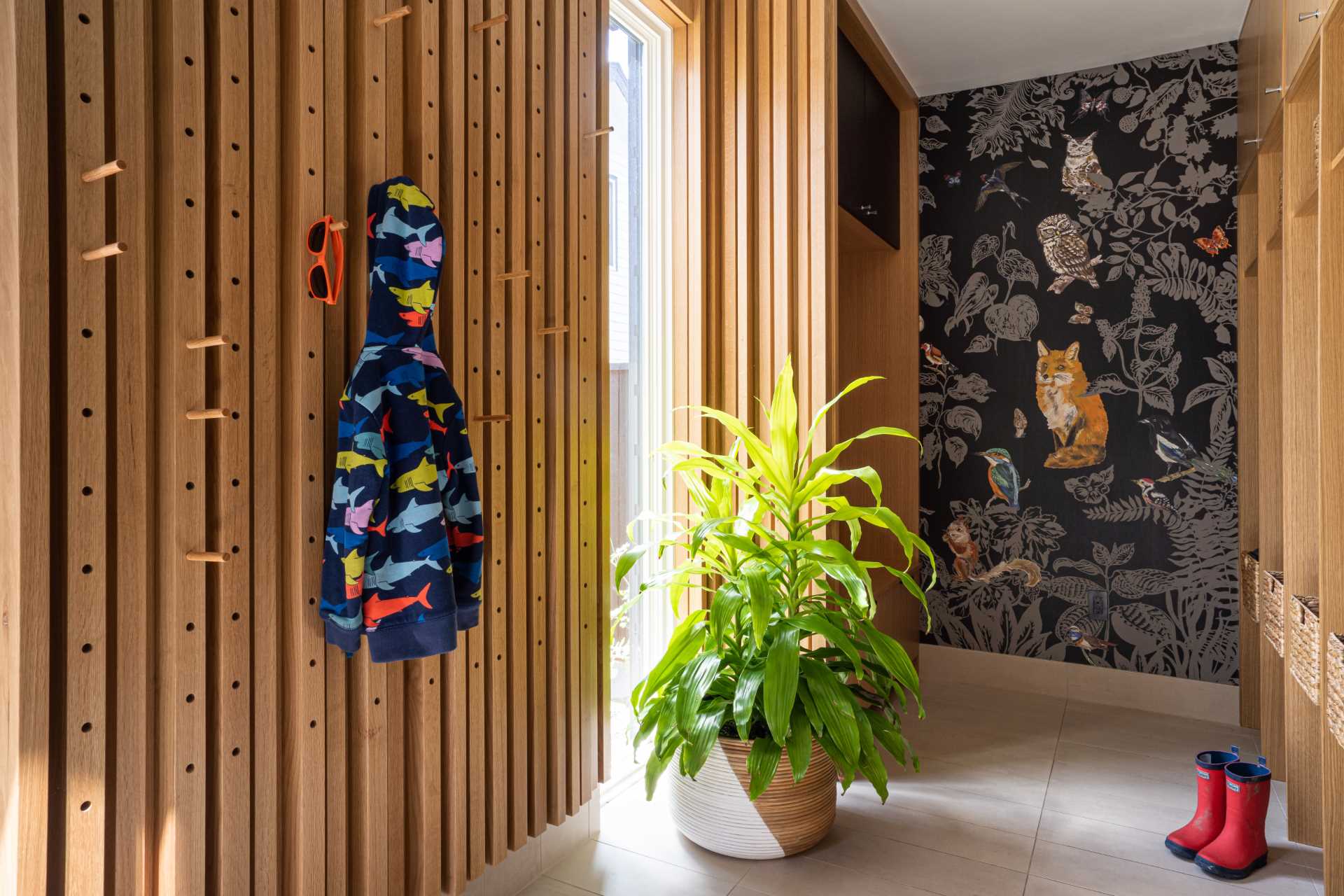 In this mudroom, custom floor-to-ceiling cabinets and shelving take up the wall, while a nature-inspired wallpaper adds an artistic element. Opposite the cabinetry is a pegboard wall that includes hooks that can be moved when needed.