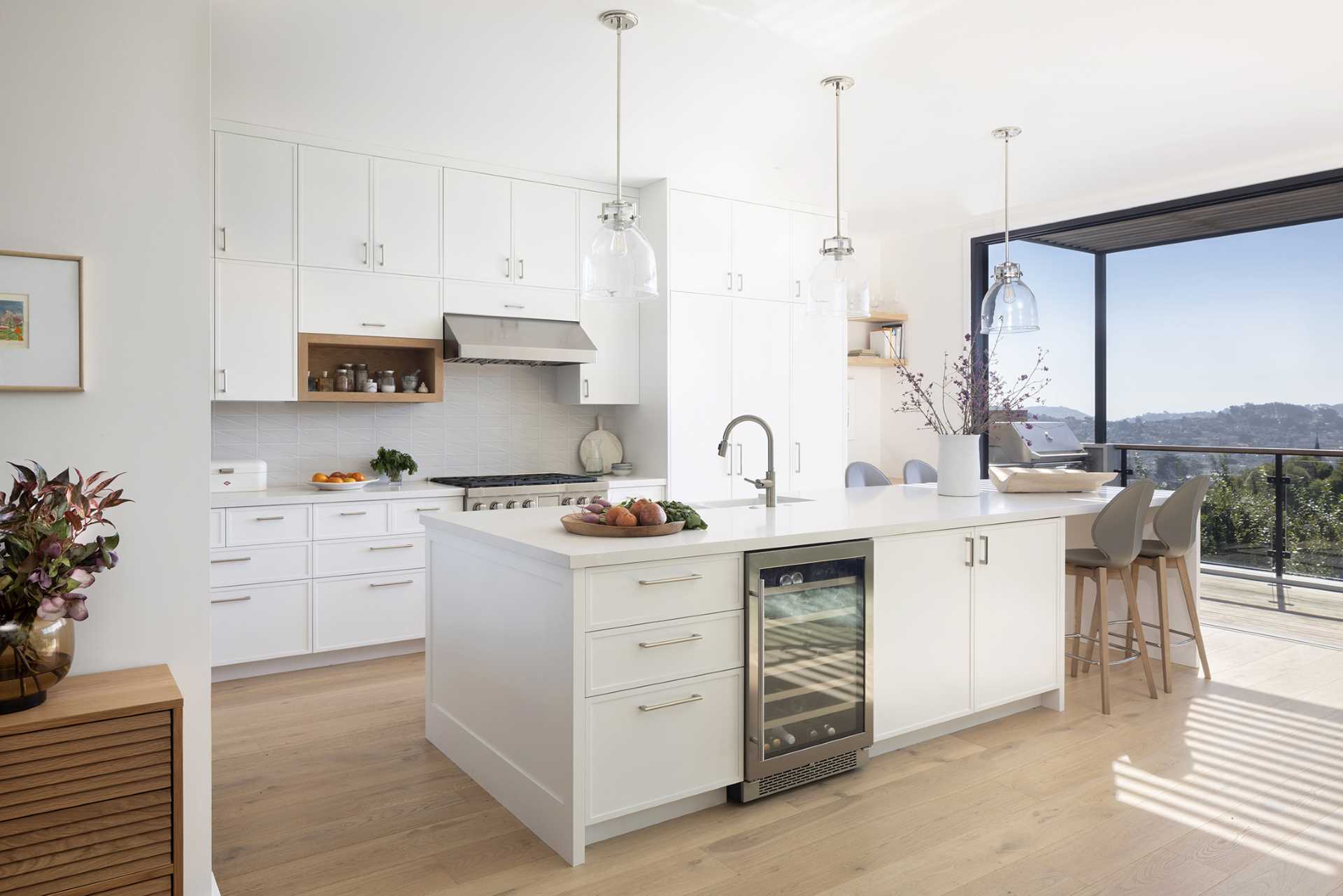 In the kitchen, white cabinets reach to the ceiling, while wood accents provide a natural touch, and a kitchen island includes a wine fridge and a place for seating. A window behind some shelves provides a glimpse of lights in the stairwell.