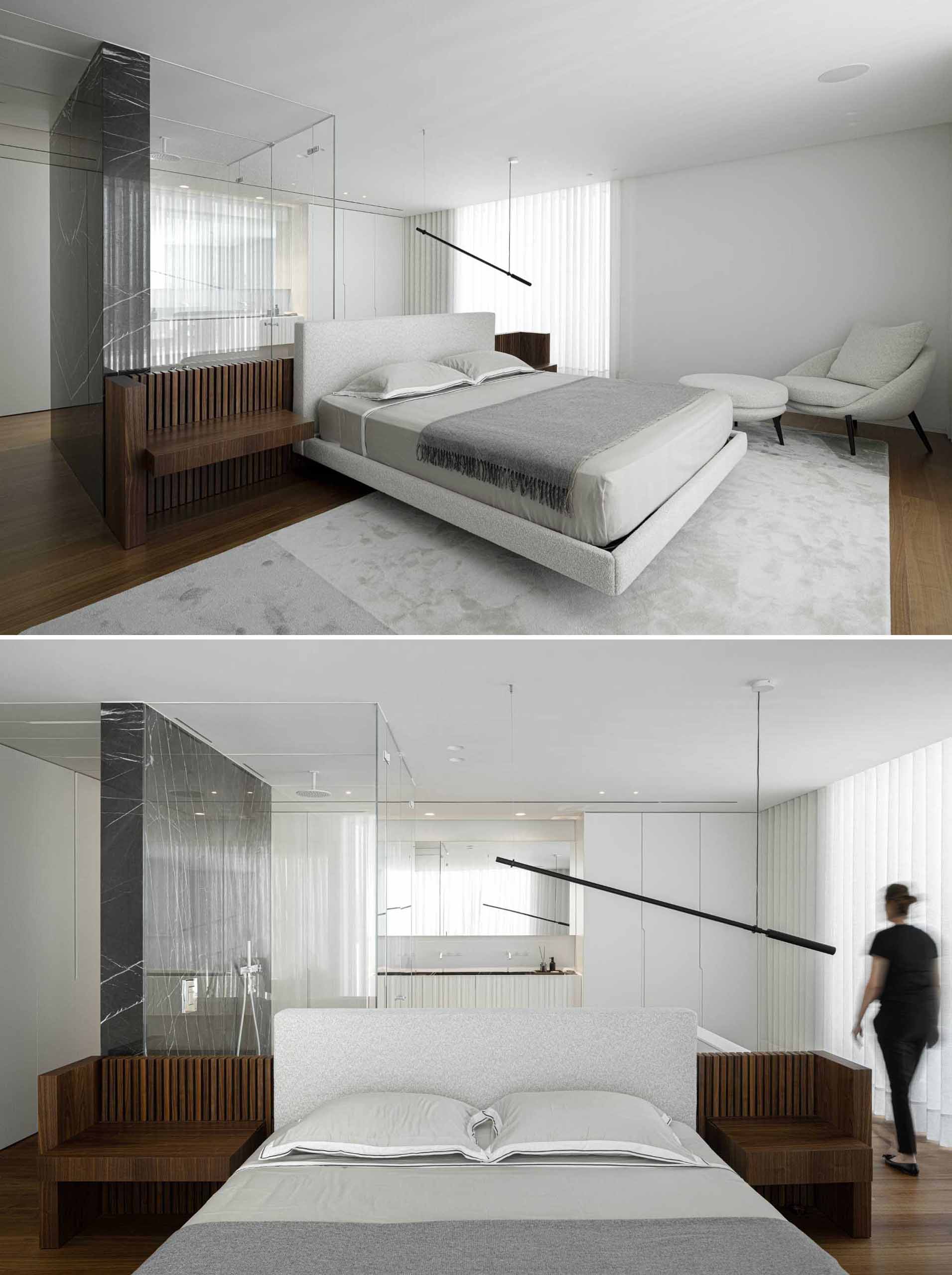 In this modern primary bedroom, the bed faces the window, while behind the bed is an en-suite bathroom with a glass-enclosed shower and a vanity that's flanked by cabinets.