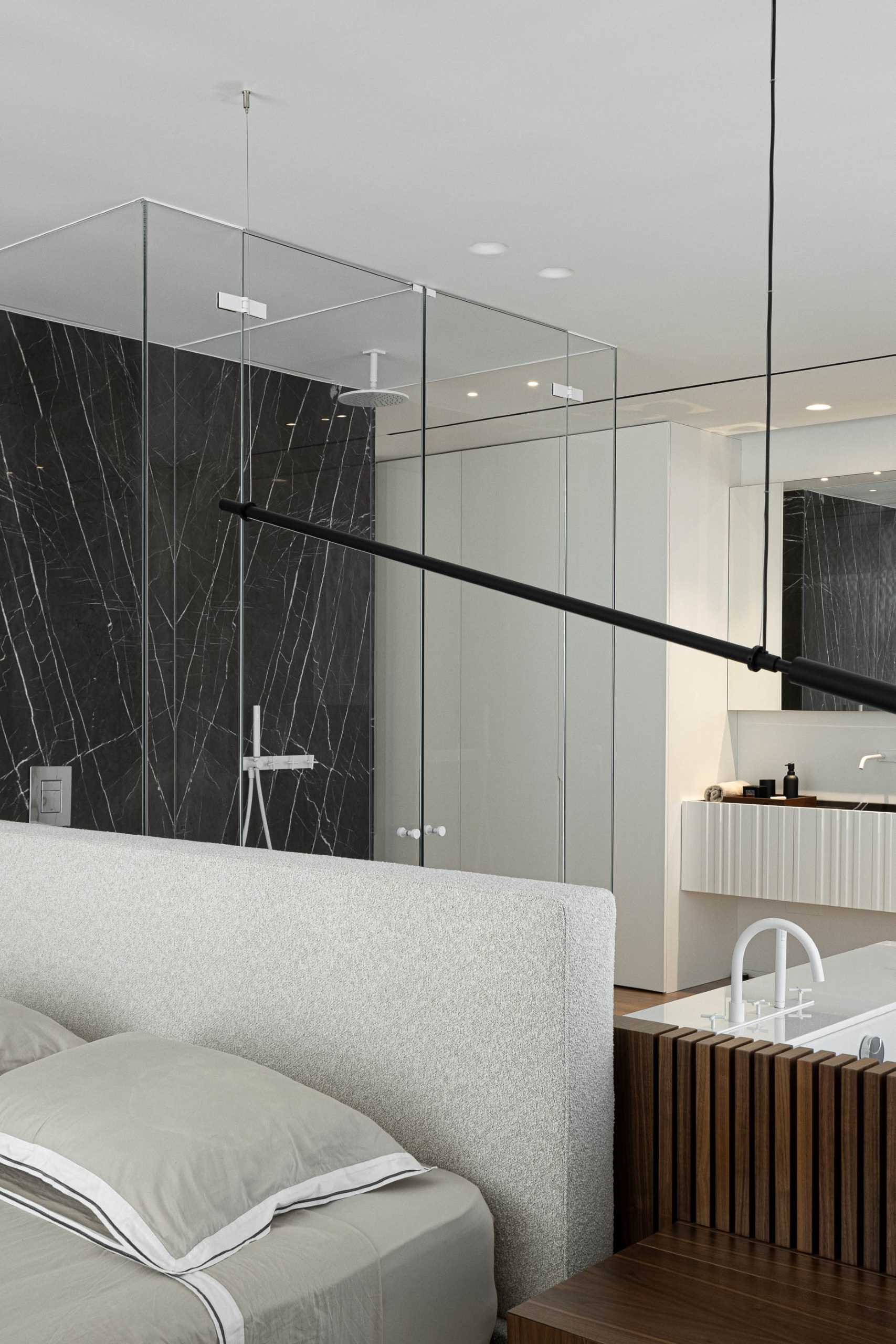 In this modern primary bedroom, the bed faces the window, while behind the bed is an en-suite bathroom with a glass-enclosed shower and a vanity that's flanked by cabinets.