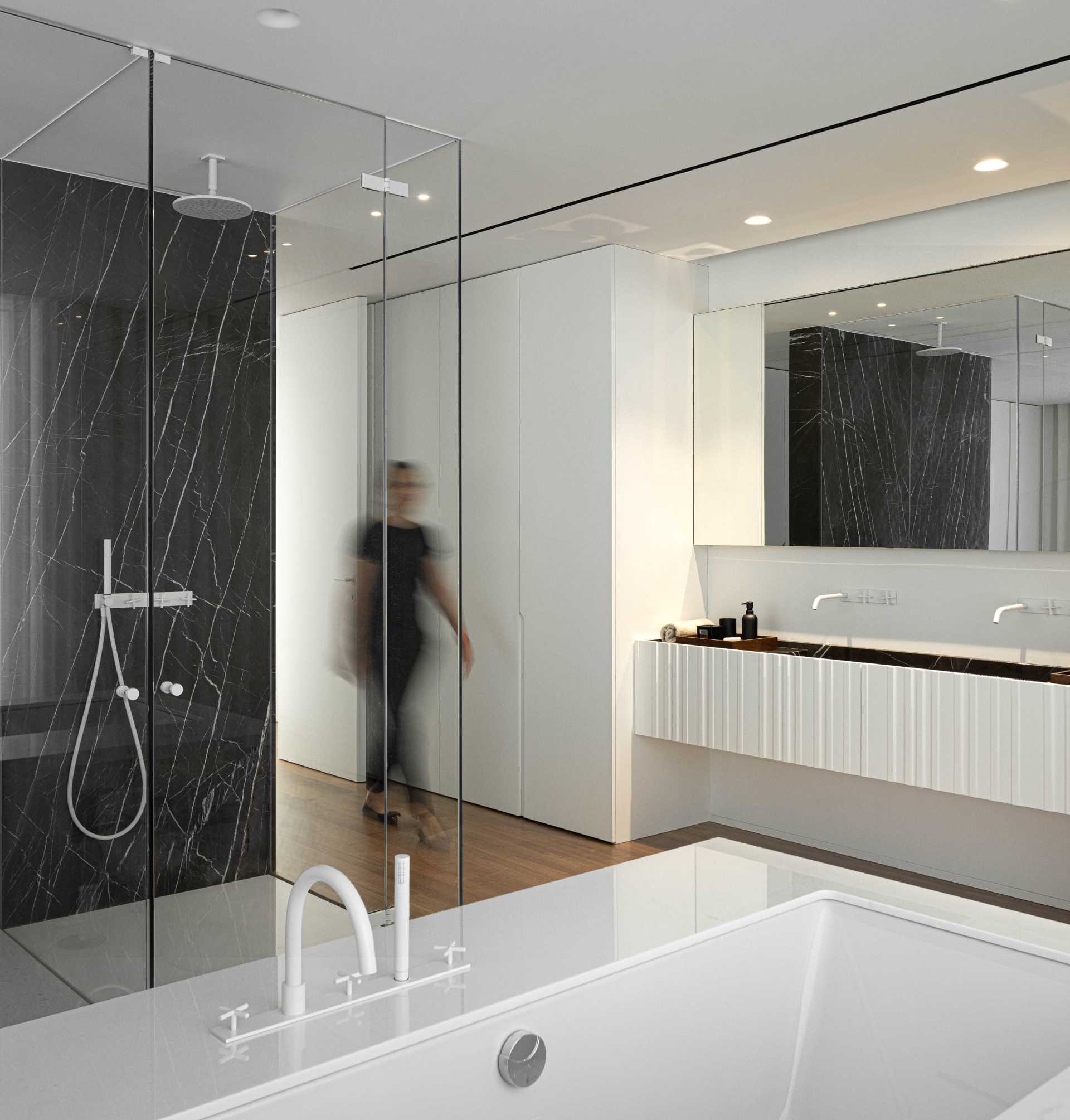 In this modern primary bedroom, the bed faces the window, while behind the bed is an en-suite bathroom with a glass-enclosed shower and a vanity that's flanked by cabinets.