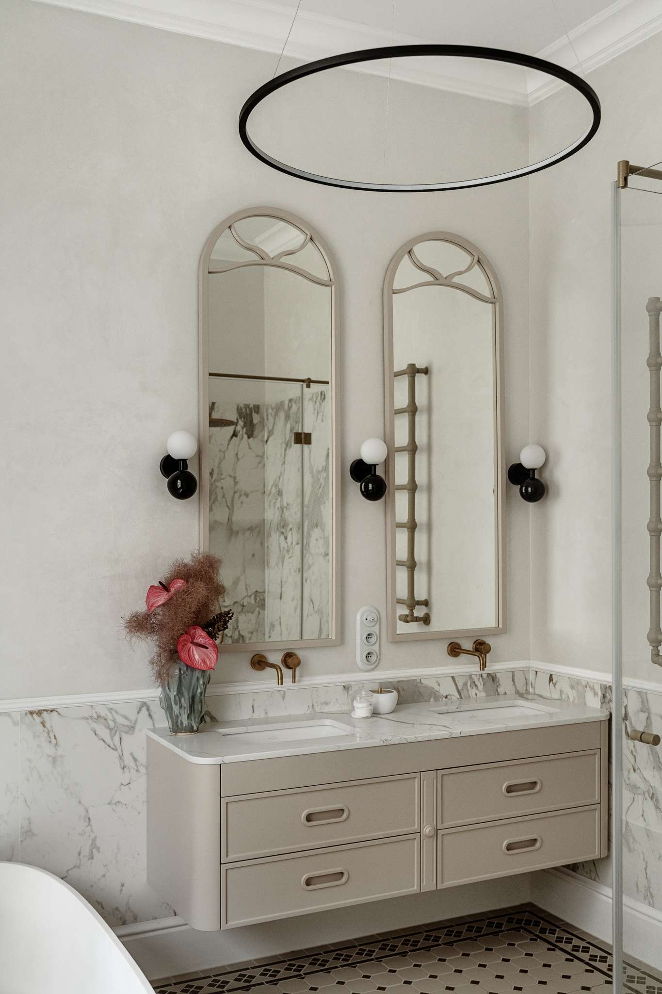 In the bathroom, classic materials and soft curves create a serene, gallery-like atmosphere. Arched mirrors nod to the apartment’s Art Nouveau heritage, while brass fixtures, decorative Ottocento plaster walls, and a floating taupe vanity bring in modern refinement. A sculptural tub with a matte finish sits beneath a sunlit window, framed by Calacatta Gold marble walls and warm metal accents.