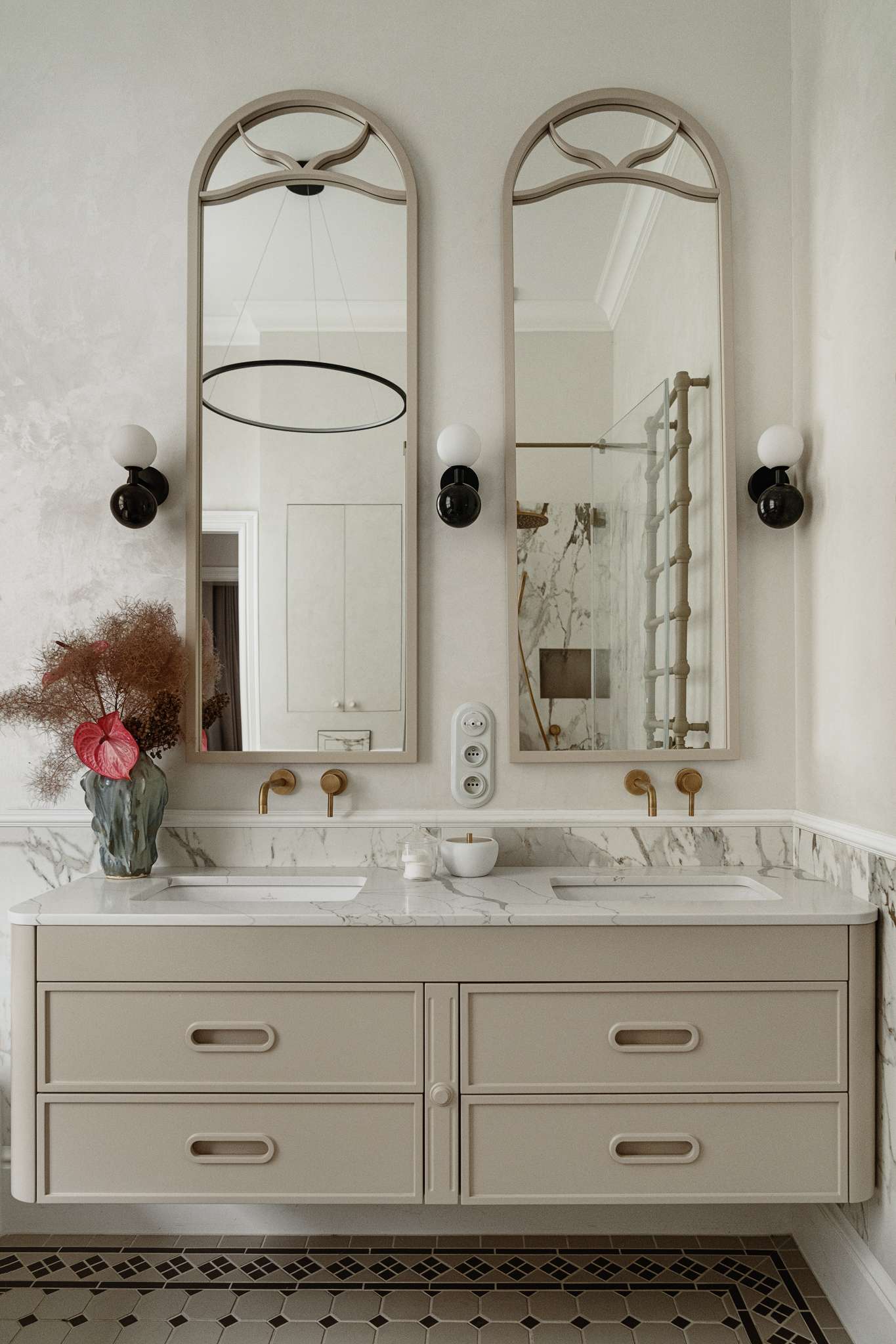 In the bathroom, classic materials and soft curves create a serene, gallery-like atmosphere. Arched mirrors nod to the apartment’s Art Nouveau heritage, while brass fixtures, decorative Ottocento plaster walls, and a floating taupe vanity bring in modern refinement. A sculptural tub with a matte finish sits beneath a sunlit window, framed by Calacatta Gold marble walls and warm metal accents.