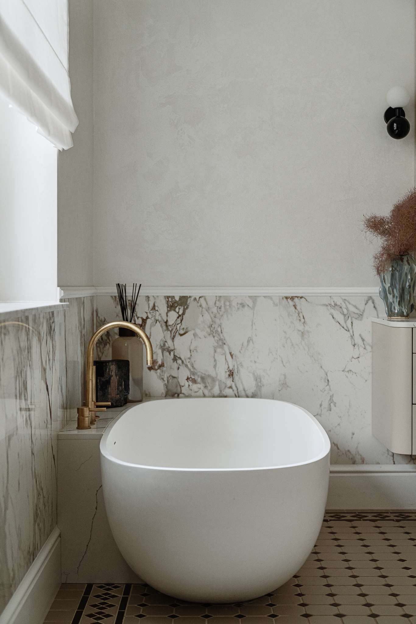 In the bathroom, classic materials and soft curves create a serene, gallery-like atmosphere. Arched mirrors nod to the apartment’s Art Nouveau heritage, while brass fixtures, decorative Ottocento plaster walls, and a floating taupe vanity bring in modern refinement. A sculptural tub with a matte finish sits beneath a sunlit window, framed by Calacatta Gold marble walls and warm metal accents.