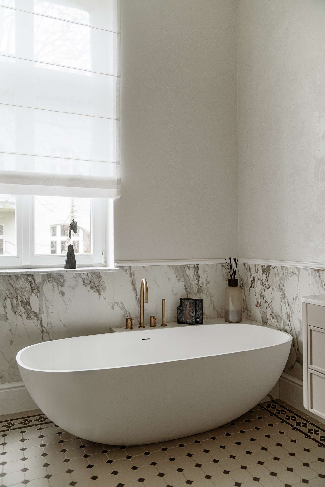 In the bathroom, classic materials and soft curves create a serene, gallery-like atmosphere. Arched mirrors nod to the apartment’s Art Nouveau heritage, while brass fixtures, decorative Ottocento plaster walls, and a floating taupe vanity bring in modern refinement. A sculptural tub with a matte finish sits beneath a sunlit window, framed by Calacatta Gold marble walls and warm metal accents.