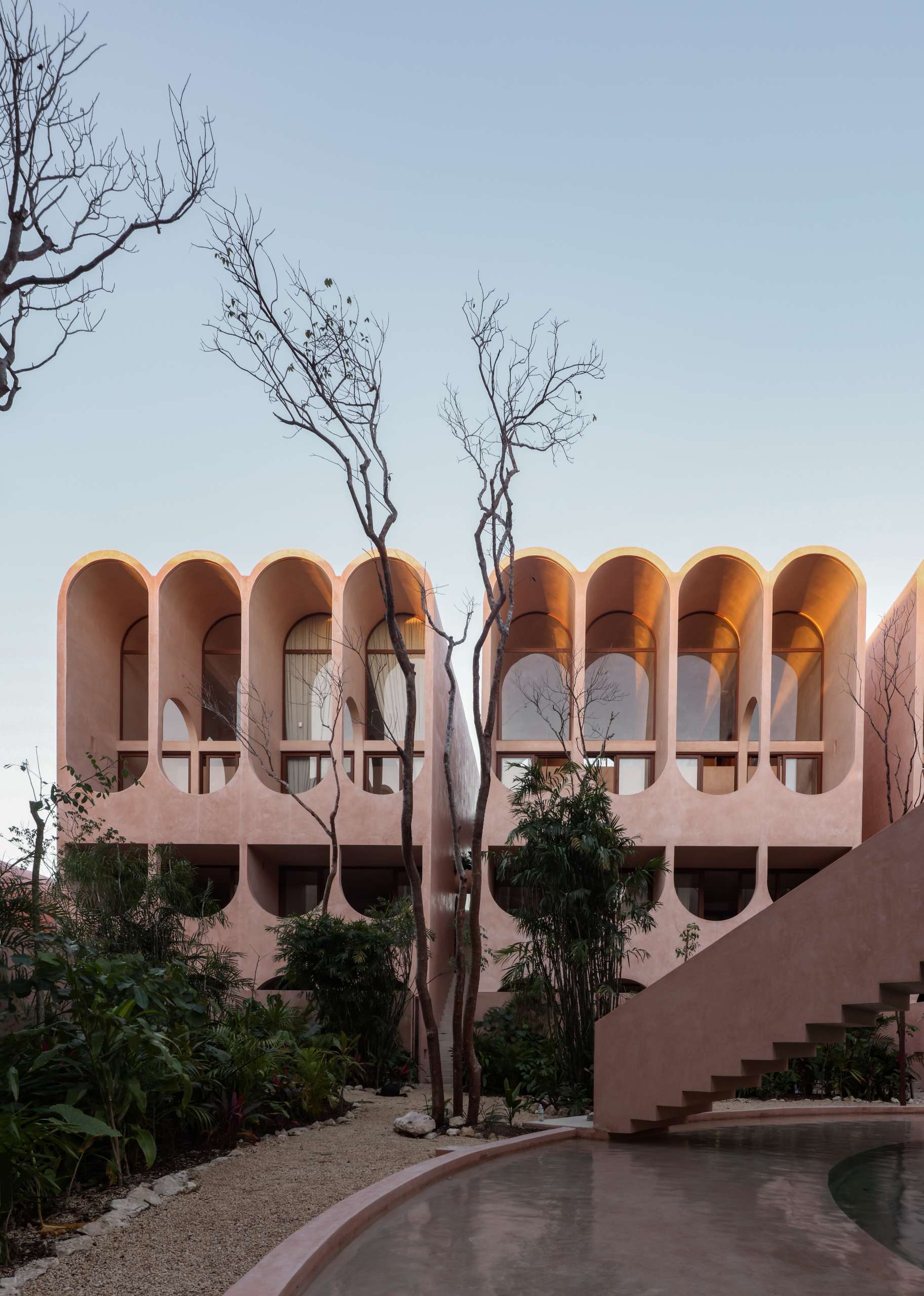 A dreamy pink facade rises behind native landscaping at this Tulum apartment complex, where arched balconies catch the golden light. The curved geometry and minimal palette create a surreal contrast between architecture and the organic jungle surroundings.