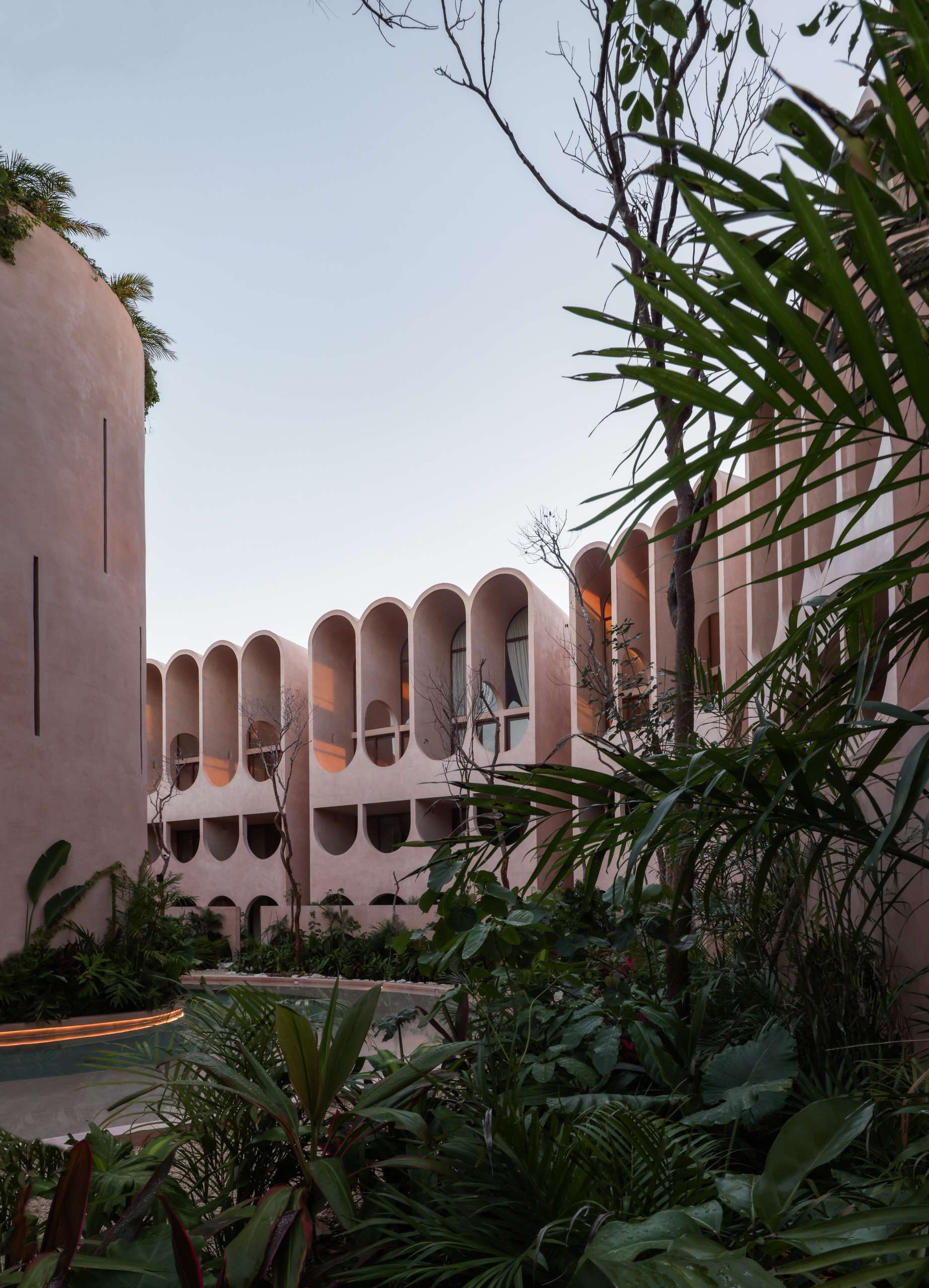 Lush greenery frames this view of a pink concrete apartment complex in Tulum, where arched openings and soft curves create a rhythm against the sky. Designed to blend with the jungle, this architecture feels both futuristic and rooted in nature.