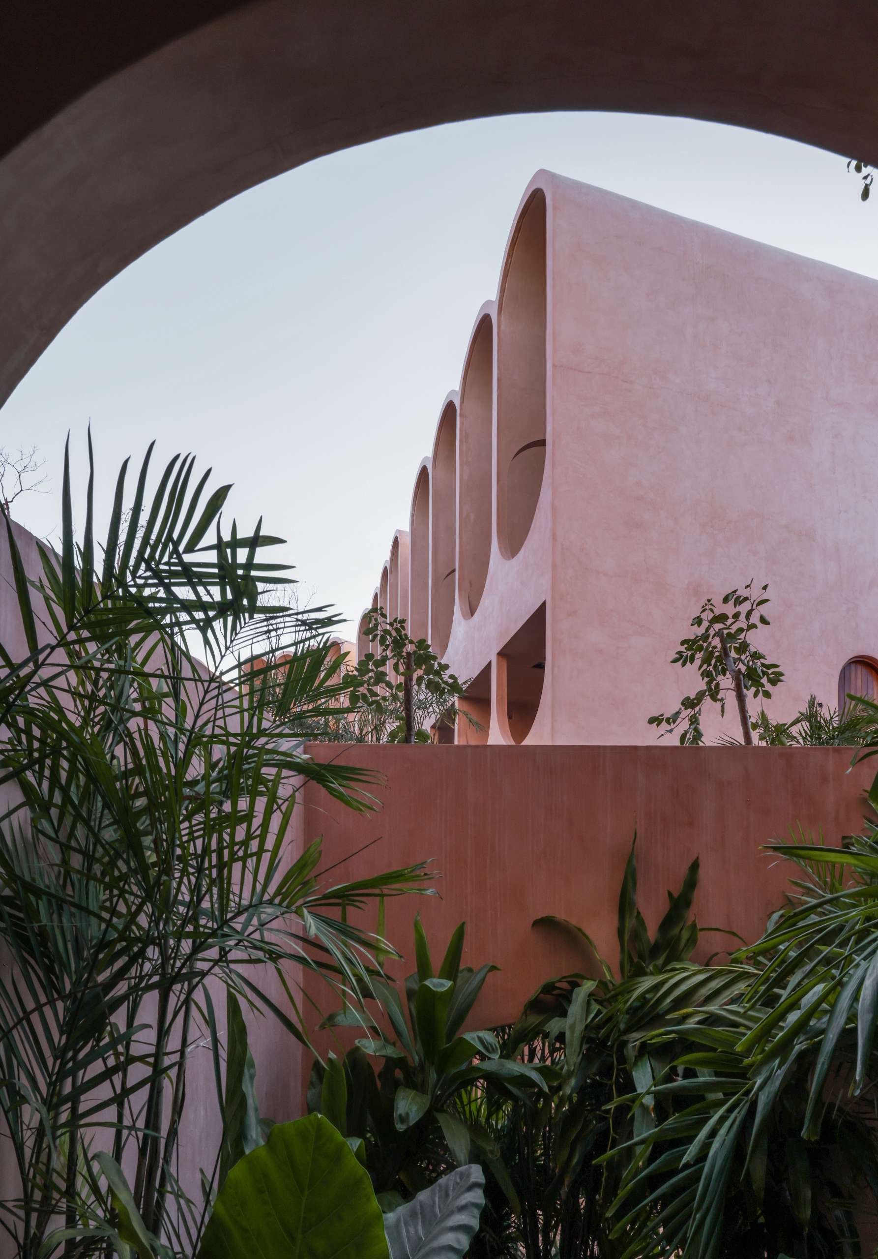 Lush greenery frames this view of a pink concrete apartment complex in Tulum, where arched openings and soft curves create a rhythm against the sky. Designed to blend with the jungle, this architecture feels both futuristic and rooted in nature.