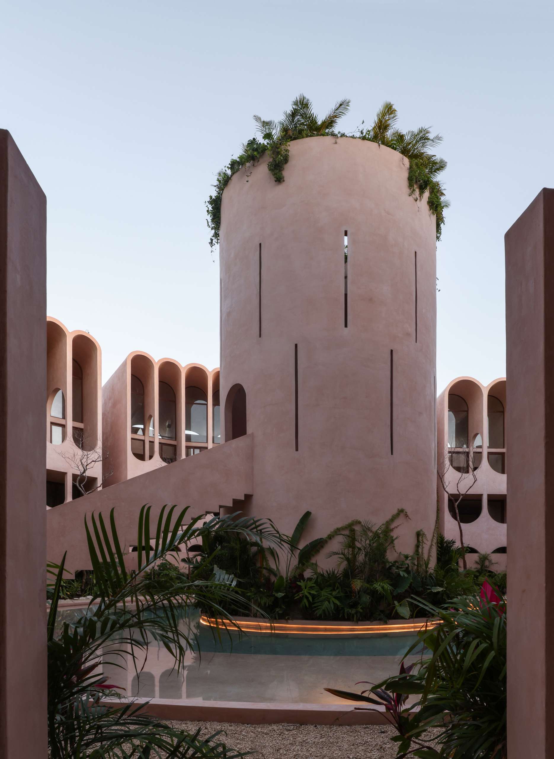 Tranquil and otherworldly, this pink concrete atrium in Tulum invites you to slow down and look up. A circular plunge pool anchors the space, while a triangular skylight lets jungle greenery spill inside. Light filters through vertical slits in the walls, casting meditative shadows across the curved interior.