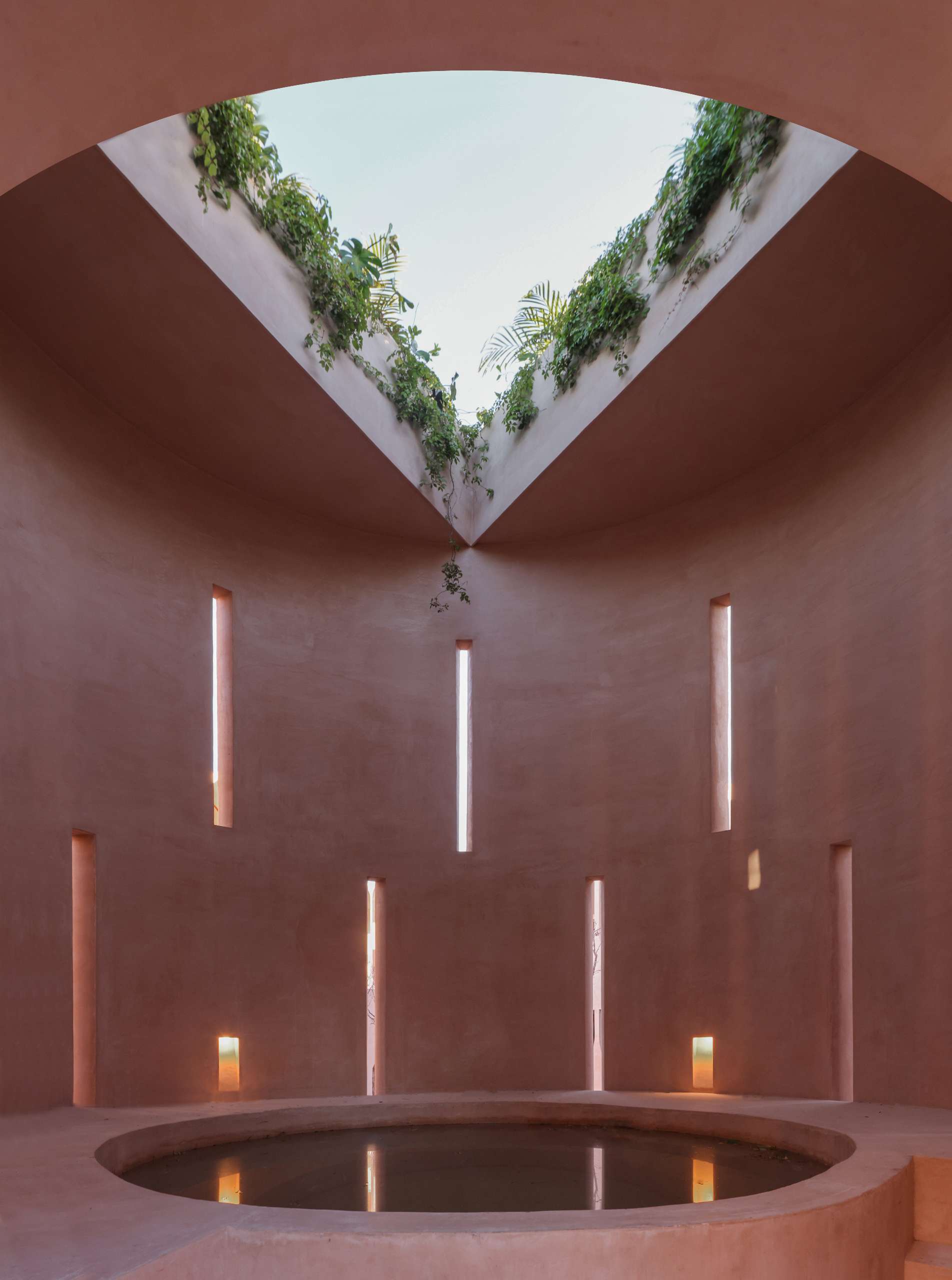 Tranquil and otherworldly, this pink concrete atrium in Tulum invites you to slow down and look up. A circular plunge pool anchors the space, while a triangular skylight lets jungle greenery spill inside. Light filters through vertical slits in the walls, casting meditative shadows across the curved interior.