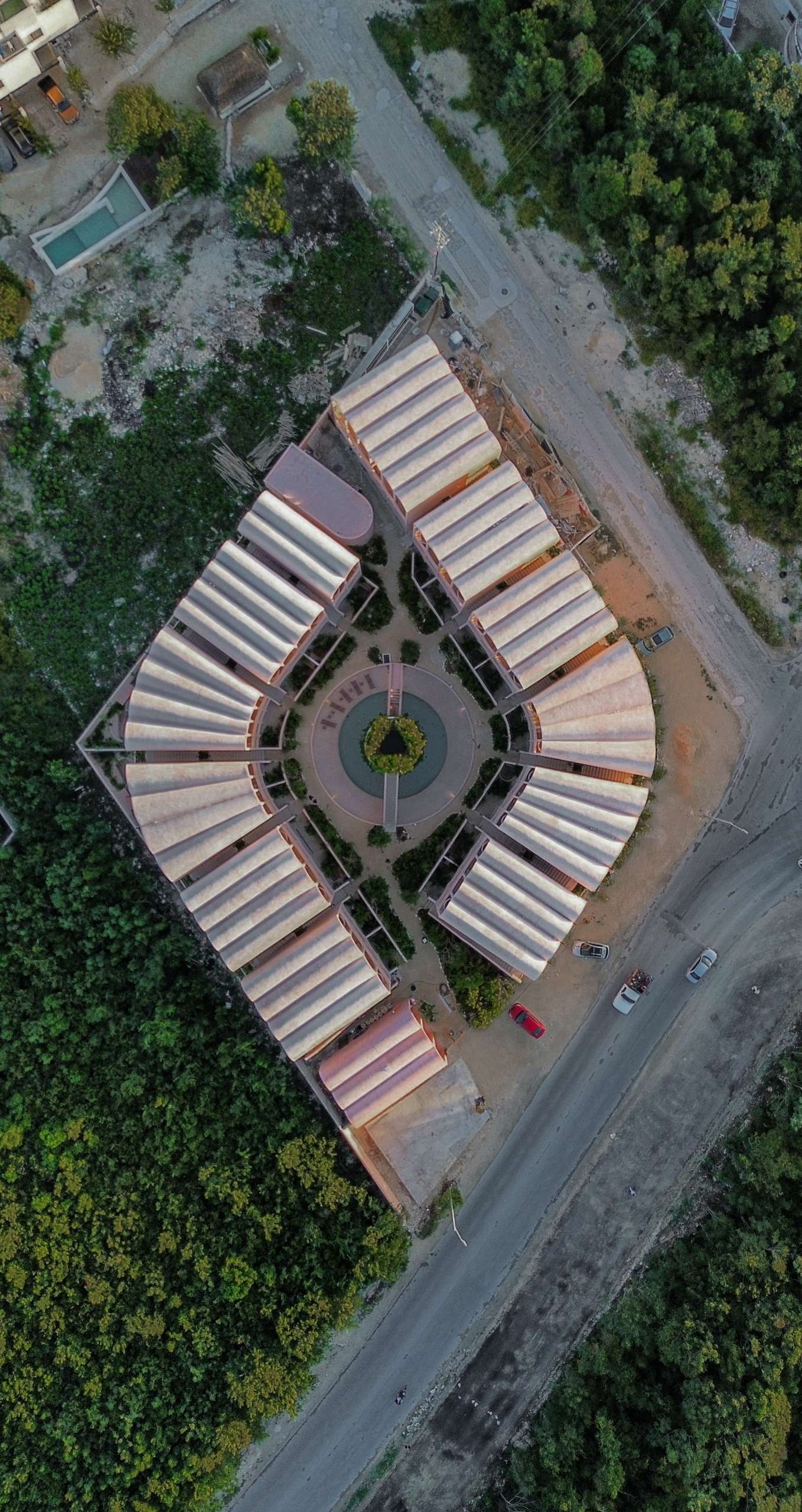 This aerial view reveals the striking symmetry of a pink-toned apartment complex nestled on the edge of the jungle in Tulum. Curved roofs and radial geometry center around a tranquil courtyard, blending bold modernism with lush tropical surroundings.