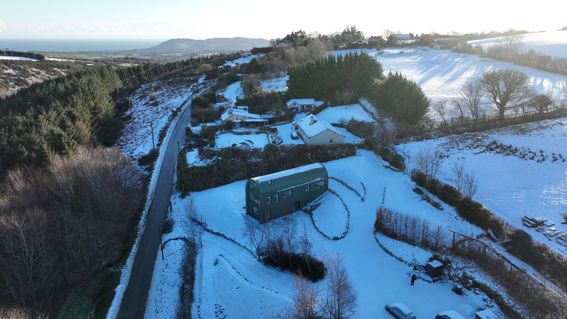 This modern home draws inspiration from the green corrugated steel sheds scattered across the Wicklow landscape, embracing their utilitarian grace while subtly elevating every detail. Its elongated form, curved roofline, and semi-submerged base follow the natural slope of the site, preserving its topographical character and minimizing excavation.