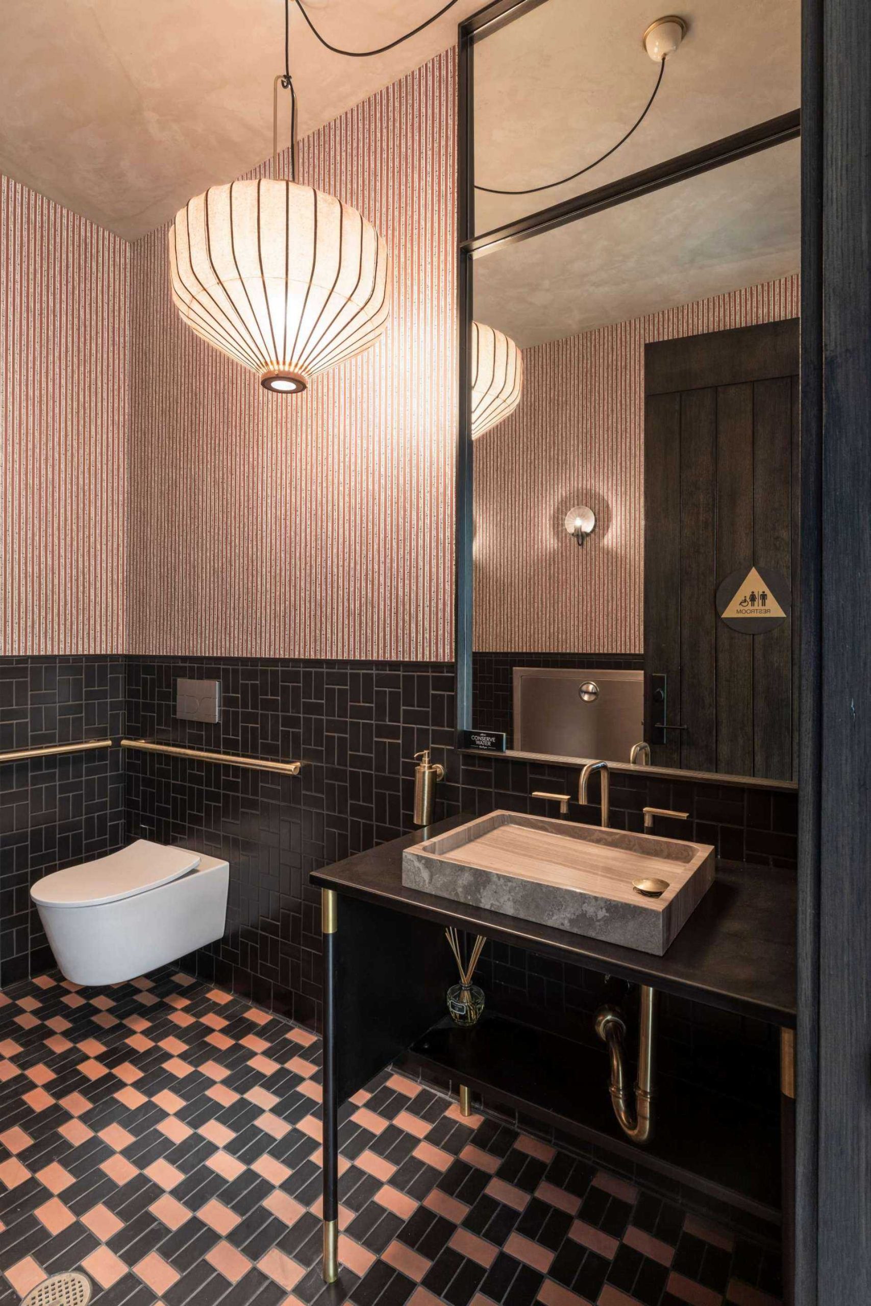 Bold and luxurious bathroom with a dramatic mix of textures and tones. Dark geometric tile and brass accents create a moody foundation, while patterned wallpaper and a lantern-style pendant light add warmth and elegance. A stone vessel sink on a black vanity gives the space a sculptural feel. This design strikes the perfect balance between vintage charm and contemporary style, ideal for boutique-inspired interiors or statement powder rooms.