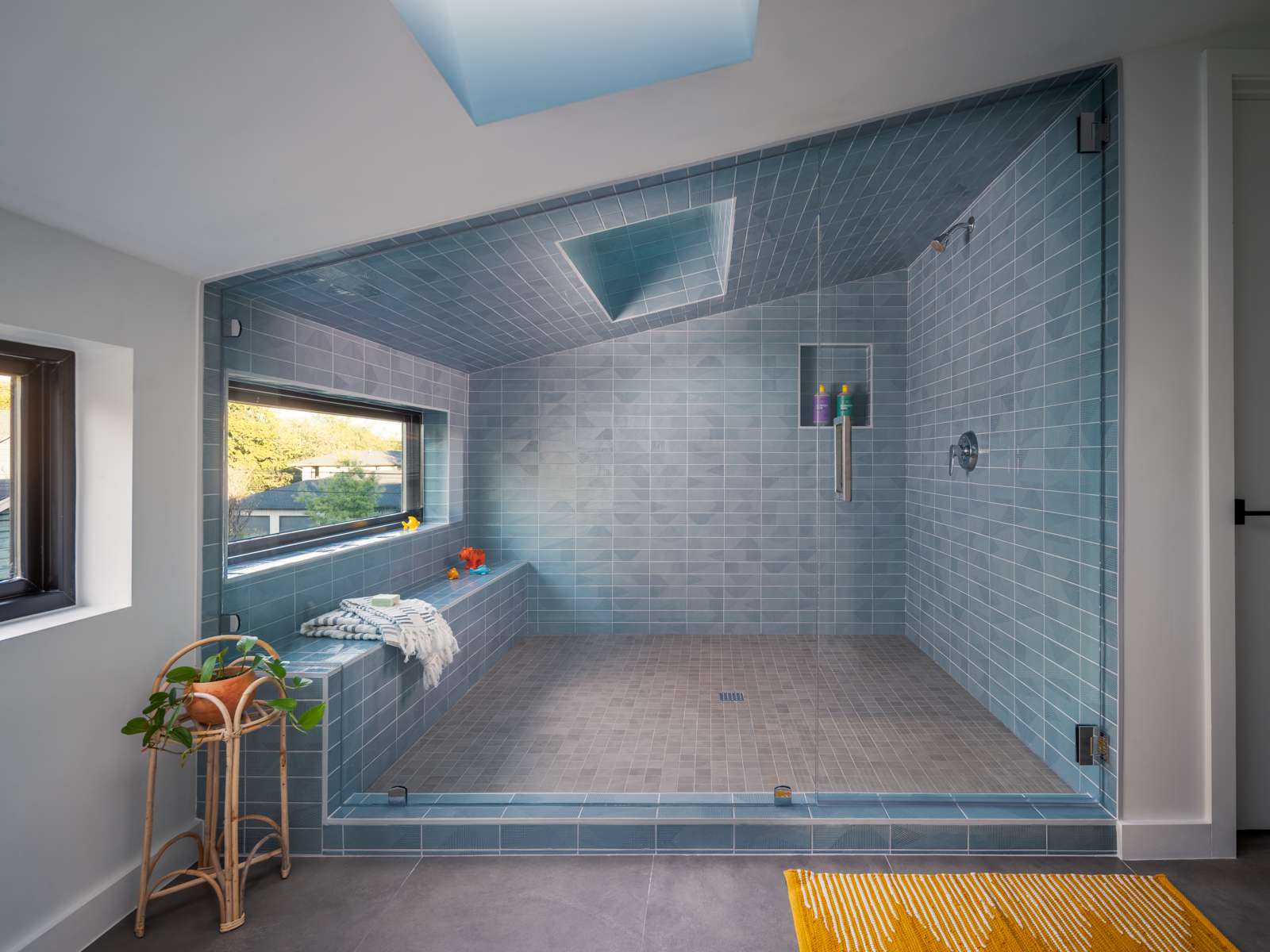 Tucked beneath an angled ceiling and surrounded by frameless glass, this expansive shower reads more like a serene spa retreat than a standard bathroom. Walls and ceiling are clad in elongated blue tiles, interrupted only by a dramatic skylight that floods the room with soft, natural light. A built-in bench runs underneath a horizontal window, while a recessed niche is neatly integrated into the tiled wall, providing a subtle storage solution.