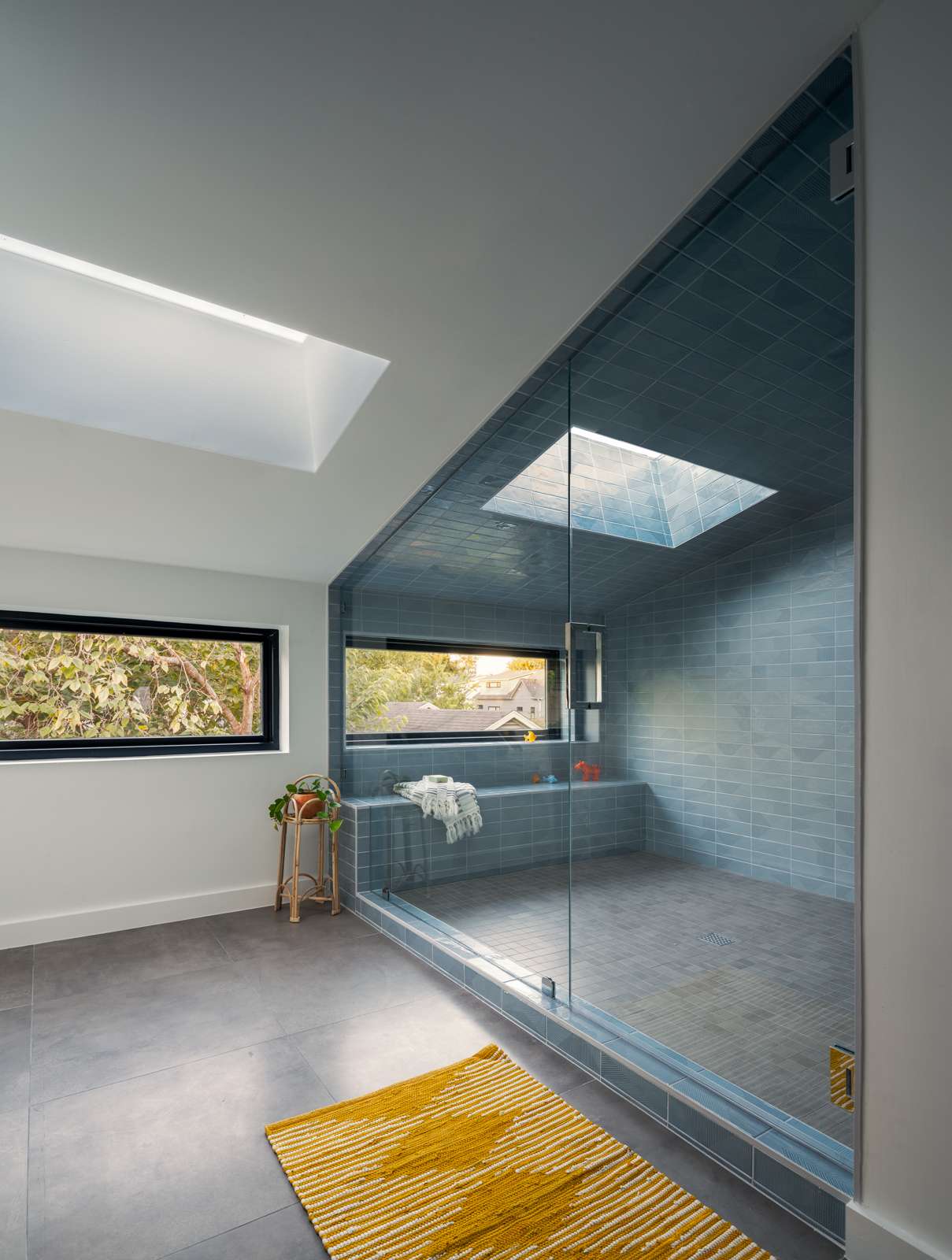 Tucked beneath an angled ceiling and surrounded by frameless glass, this expansive shower reads more like a serene spa retreat than a standard bathroom. Walls and ceiling are clad in elongated blue tiles, interrupted only by a dramatic skylight that floods the room with soft, natural light. A built-in bench runs underneath a horizontal window, while a recessed niche is neatly integrated into the tiled wall, providing a subtle storage solution.