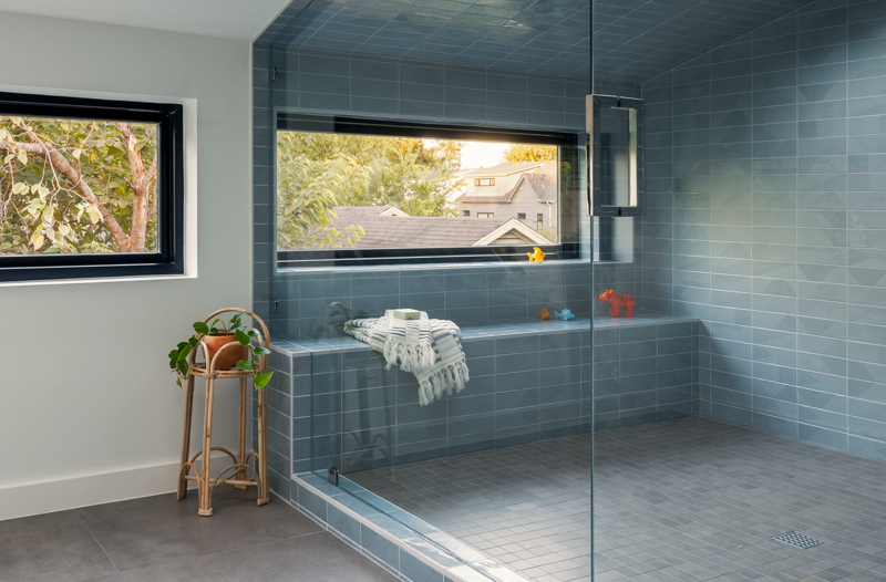 Tucked beneath an angled ceiling and surrounded by frameless glass, this expansive shower reads more like a serene spa retreat than a standard bathroom. Walls and ceiling are clad in elongated blue tiles, interrupted only by a dramatic skylight that floods the room with soft, natural light. A built-in bench runs underneath a horizontal window, while a recessed niche is neatly integrated into the tiled wall, providing a subtle storage solution.