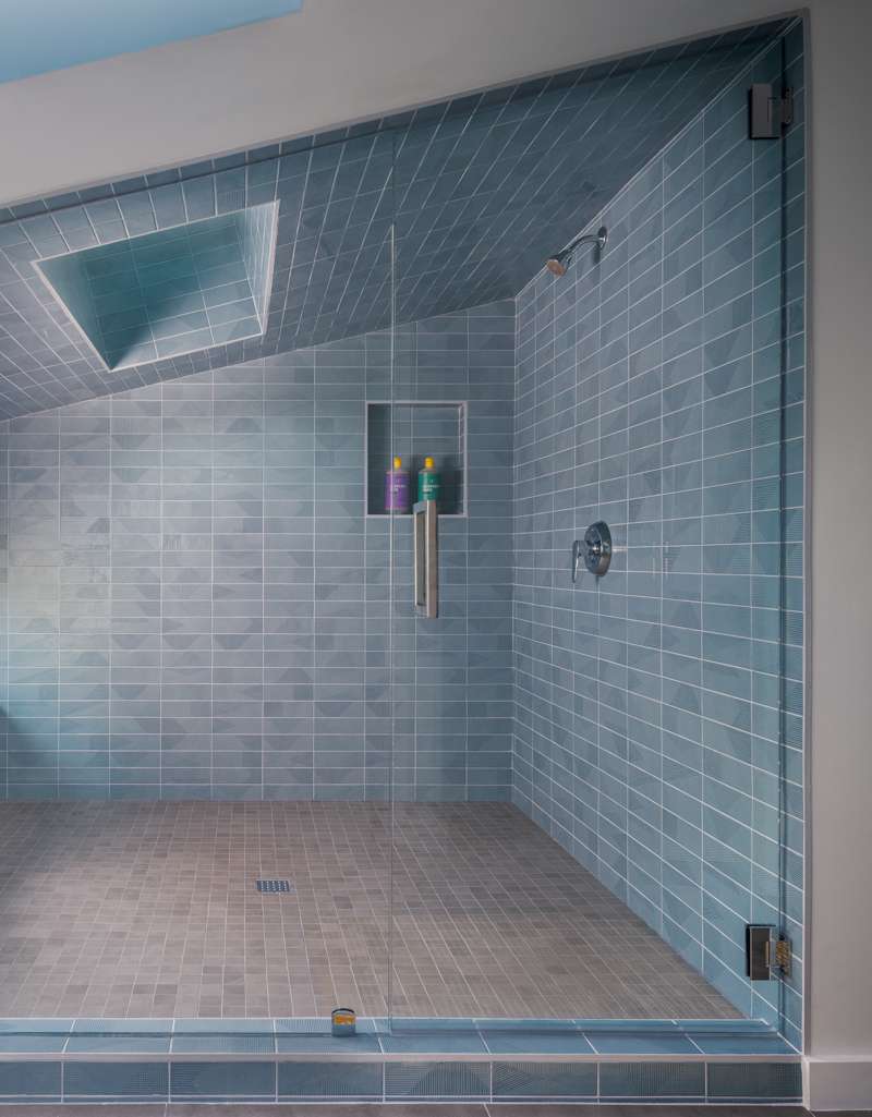 Tucked beneath an angled ceiling and surrounded by frameless glass, this expansive shower reads more like a serene spa retreat than a standard bathroom. Walls and ceiling are clad in elongated blue tiles, interrupted only by a dramatic skylight that floods the room with soft, natural light. A built-in bench runs underneath a horizontal window, while a recessed niche is neatly integrated into the tiled wall, providing a subtle storage solution.