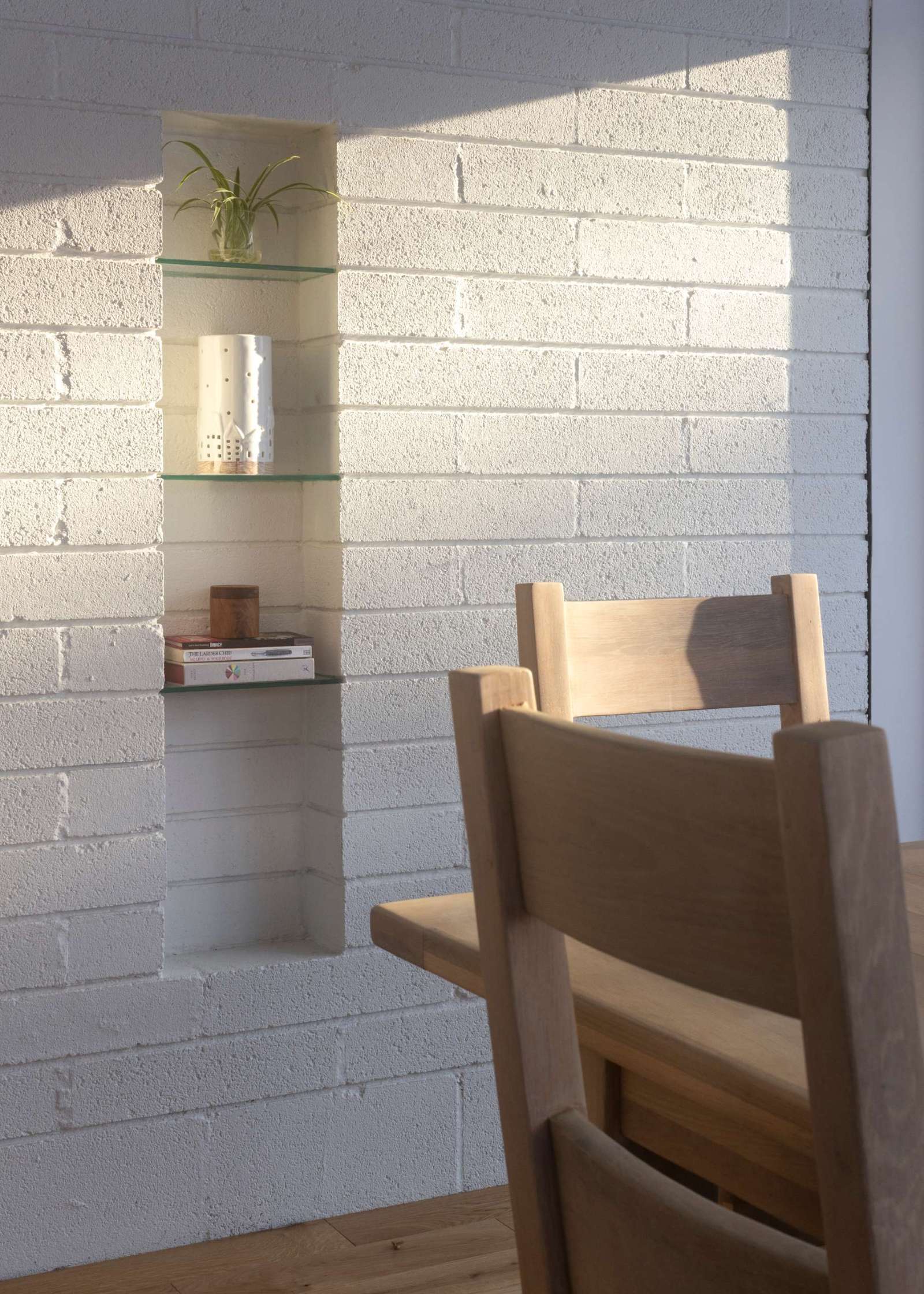In this dining area, simple and stylish wall niche shelving has set into white-painted brick for a clean look. Glass shelves keep the display light and airy, perfect for showcasing ceramics, books, or small plants. Paired with warm wood furniture and soft morning light, this built-in feature adds character and function.