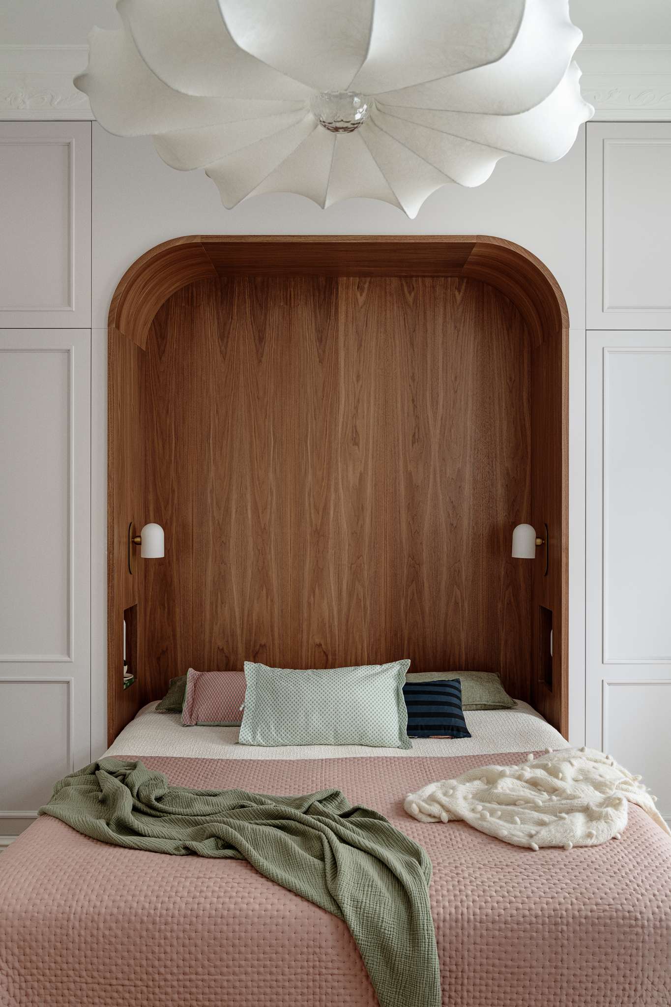 The bedroom blends warmth, softness, and subtle historic detail. A custom walnut niche wraps the bed in a cozy curve, with built-in lights and shelving for quiet functionality. Pale grey wardrobes with brass knobs frame the space, while blush bedding, sage accents, and a sculptural pendant light add layers of texture and calm.