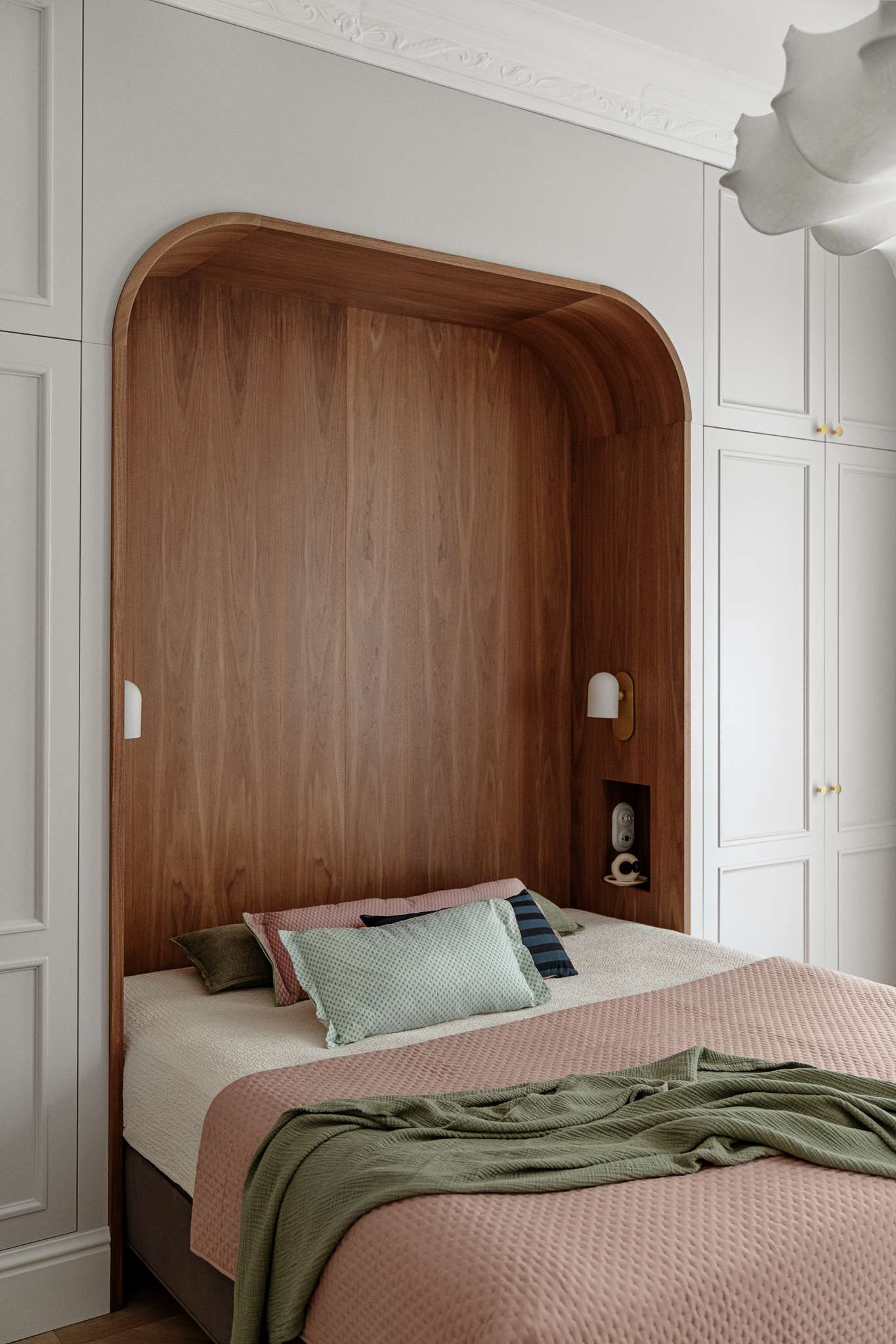 The bedroom blends warmth, softness, and subtle historic detail. A custom walnut niche wraps the bed in a cozy curve, with built-in lights and shelving for quiet functionality. Pale grey wardrobes with brass knobs frame the space, while blush bedding, sage accents, and a sculptural pendant light add layers of texture and calm.