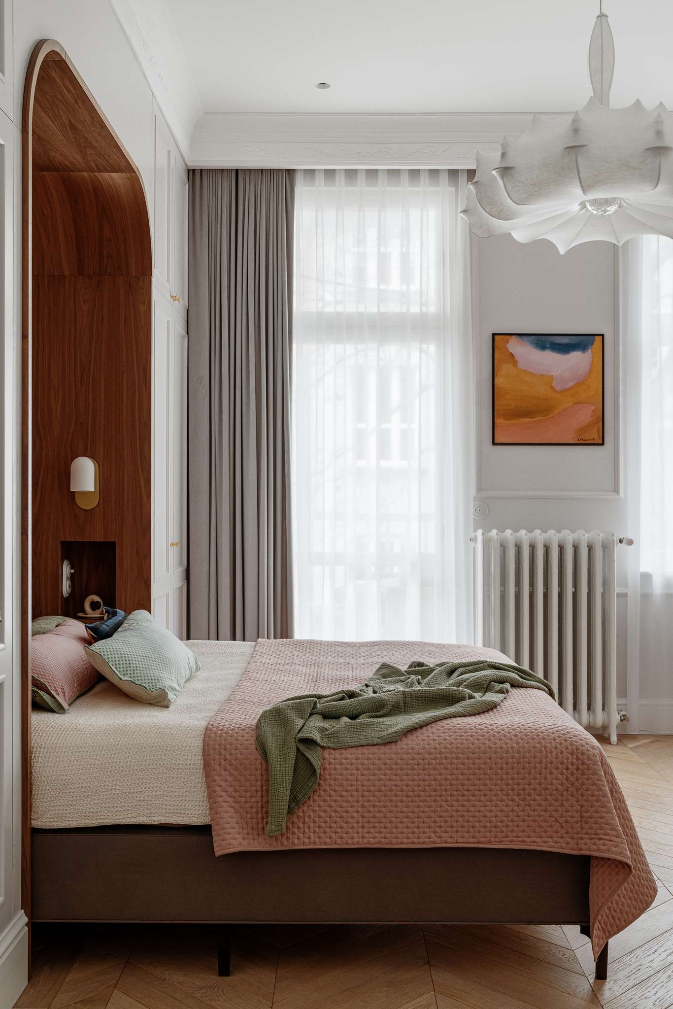 The bedroom blends warmth, softness, and subtle historic detail. A custom walnut niche wraps the bed in a cozy curve, with built-in lights and shelving for quiet functionality. Pale grey wardrobes with brass knobs frame the space, while blush bedding, sage accents, and a sculptural pendant light add layers of texture and calm.
