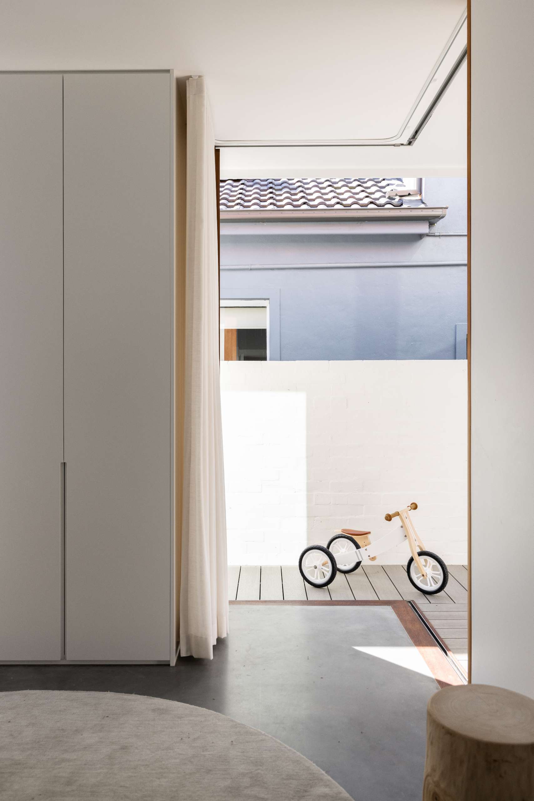 Corner sliding doors open this minimalist playroom to a private deck, while sheer curtains and built-in cabinetry keep the space bright and clutter-free. A round rug softens the polished concrete floor for a cozy play area.