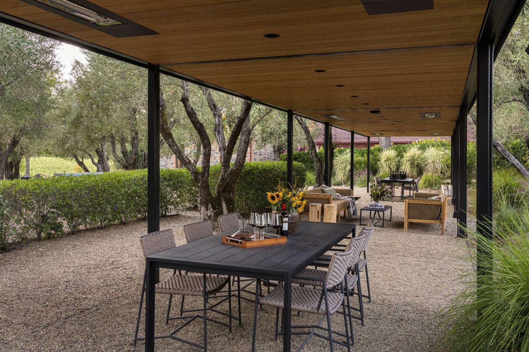 Under a wood-lined pergola, this outdoor dining area continues the tasting room’s refined yet relaxed aesthetic. Black steel framing and woven chairs echo the interior’s textures, while trees and gravel ground the space in its natural Napa setting.