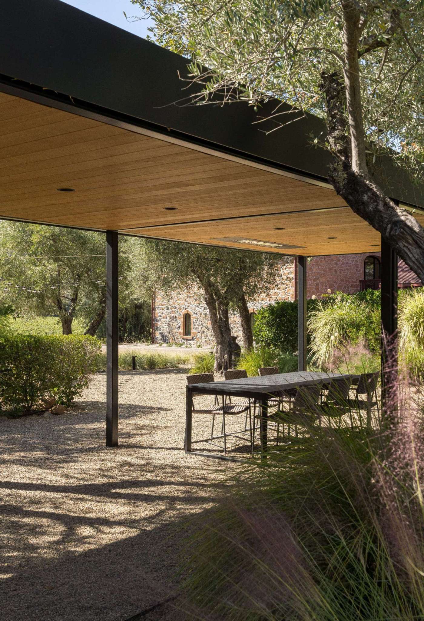 Under a wood-lined pergola, this outdoor dining area continues the tasting room’s refined yet relaxed aesthetic. Black steel framing and woven chairs echo the interior’s textures, while trees and gravel ground the space in its natural Napa setting.