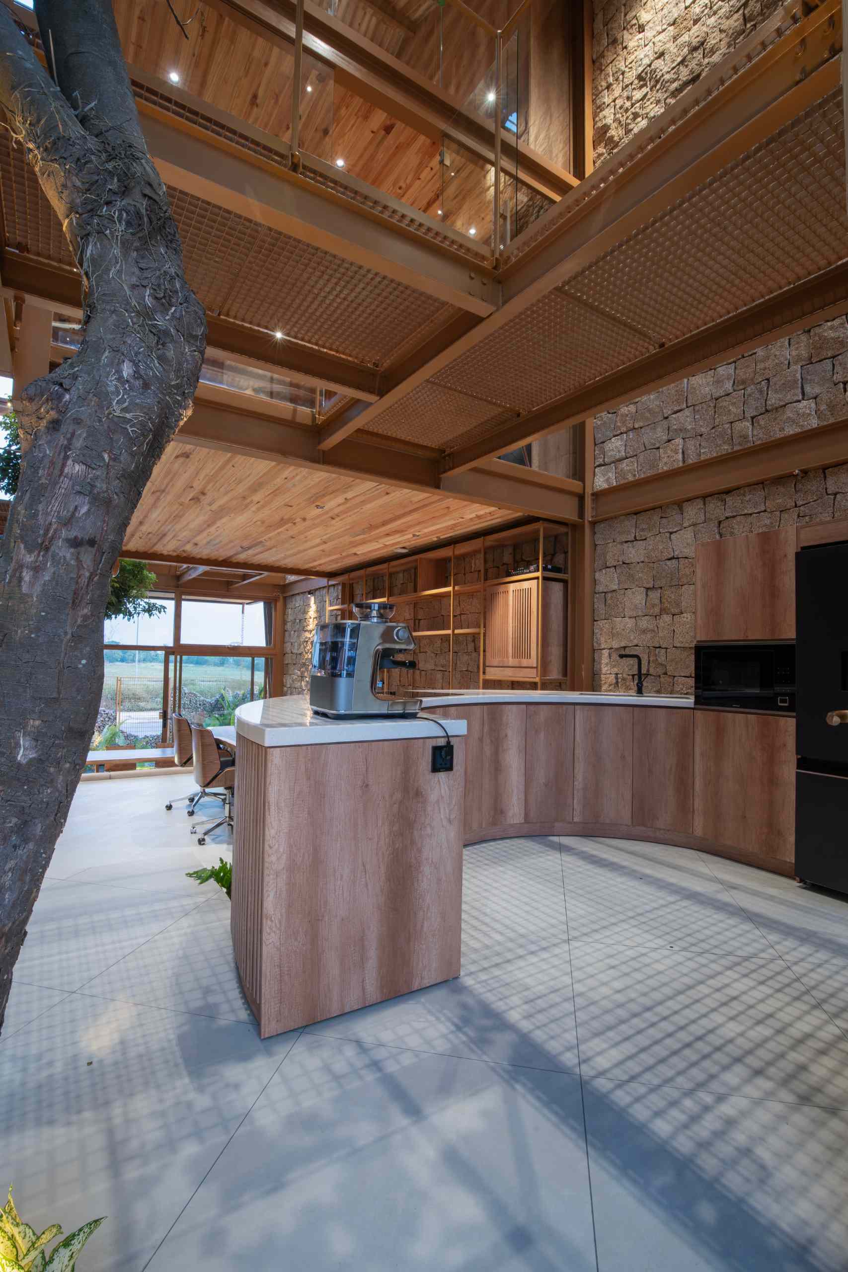 A curved wooden counter anchors the kitchen. Trees grow right through the floor, blurring the line between indoors and out, while sunlight filters through the mesh walkways above, casting a soft grid of shadows.