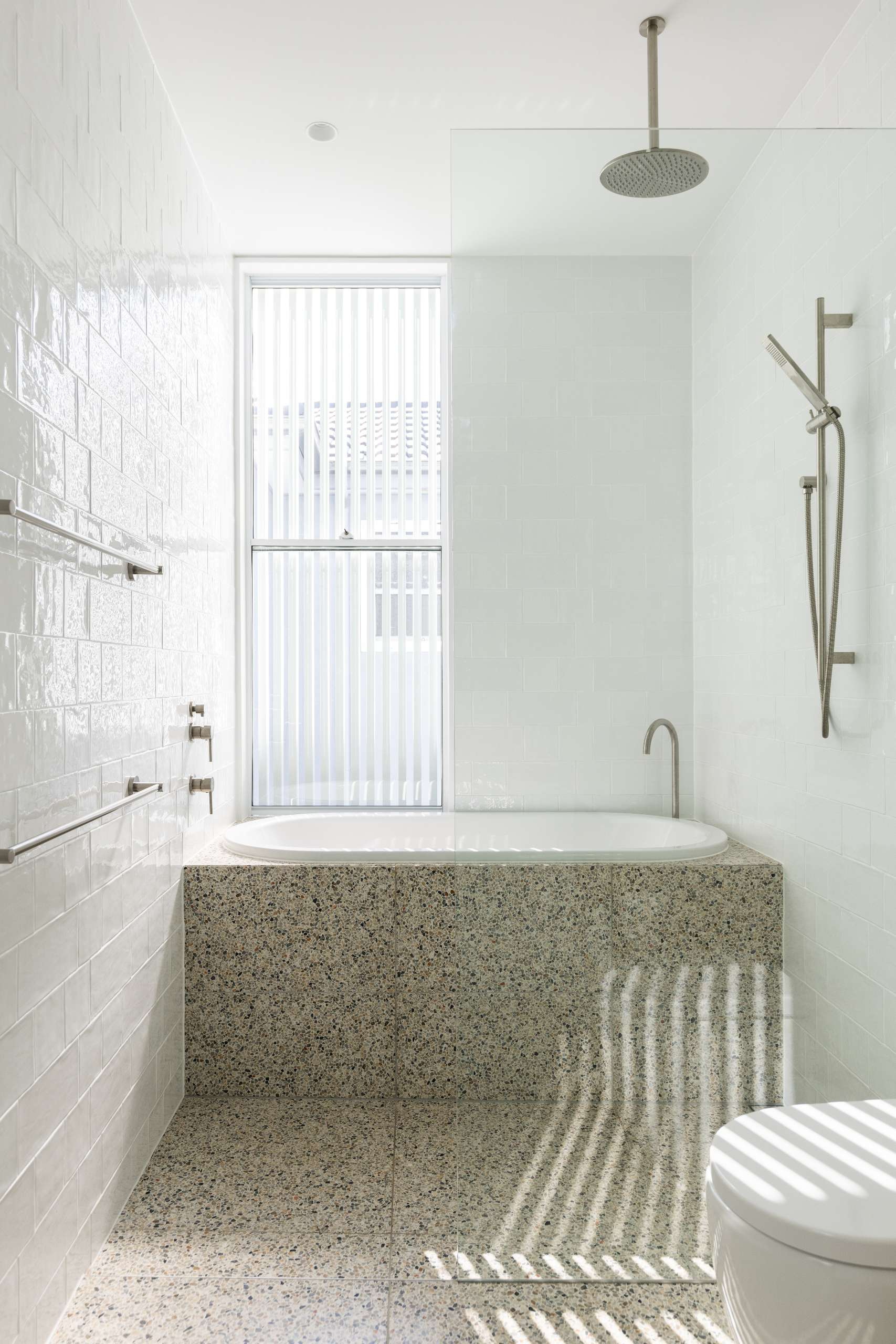 A clean and calming family bathroom where terrazzo floors and a built-in bathtub bring warmth and durability. The rainfall shower, soft white tiles, and filtered natural light create a minimalist retreat that’s as practical as it is peaceful.