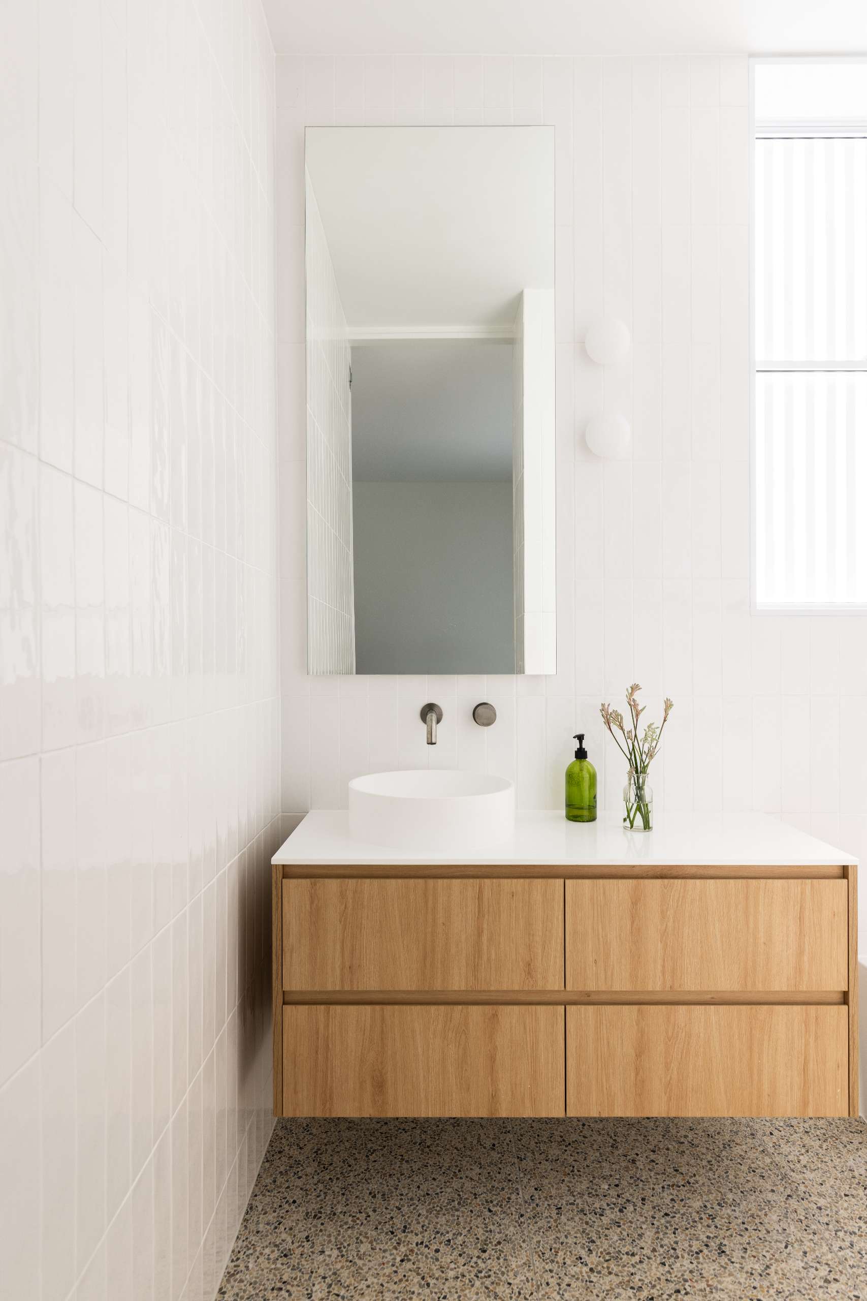 A serene ensuite moment with a minimalist wood vanity, round vessel sink, and wall-mounted tapware. Glossy white tiles and soft natural light amplify the calm, while a simple sprig in a glass jar brings a quiet, organic touch.