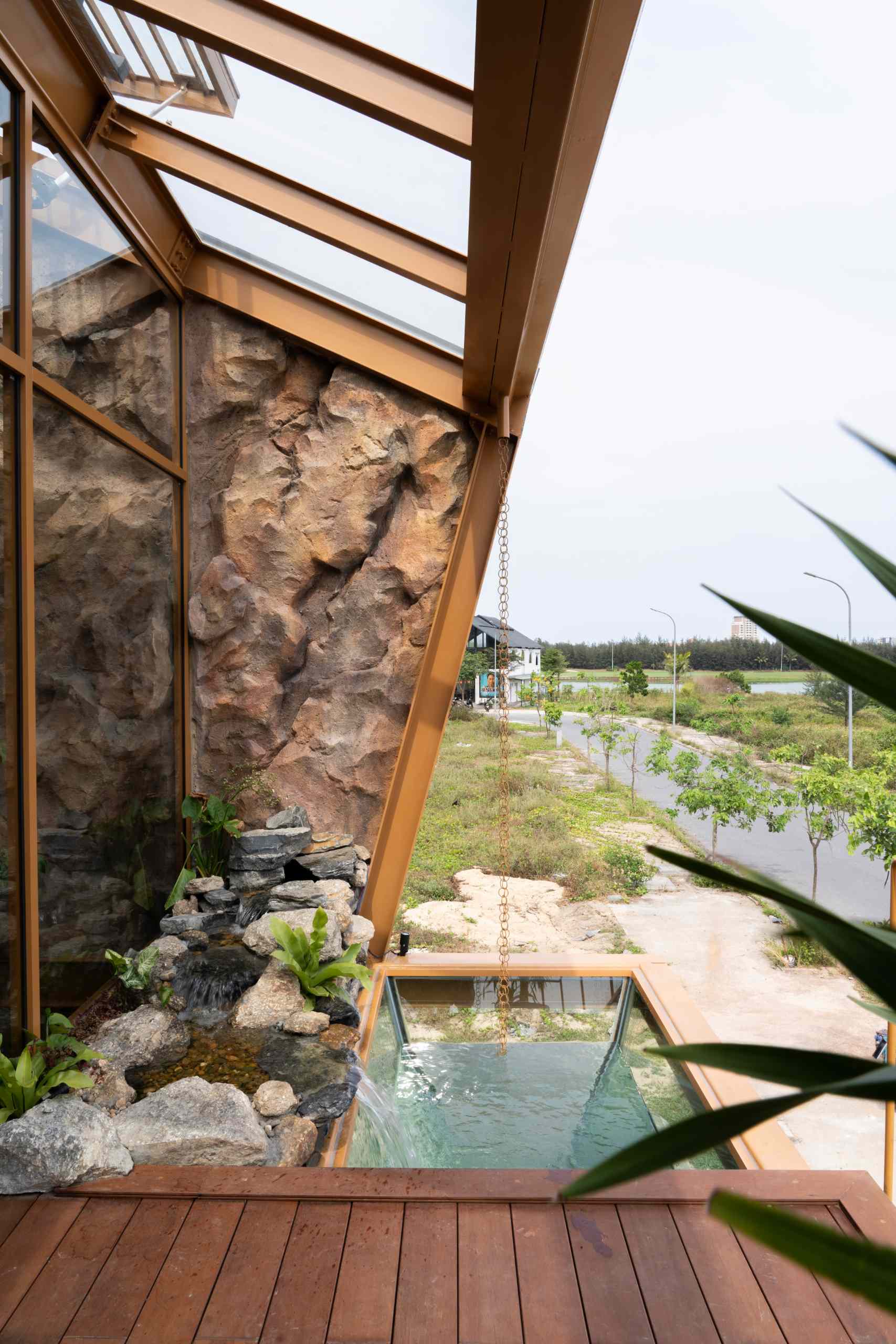 A glass volume suspended above a pond creates a striking waterfall that cools the space and adds a calming sound. Surrounded by tropical plants, natural stone, and a textured gabion wall, the garden feels like a small, living ecosystem built into the heart of a modern home.