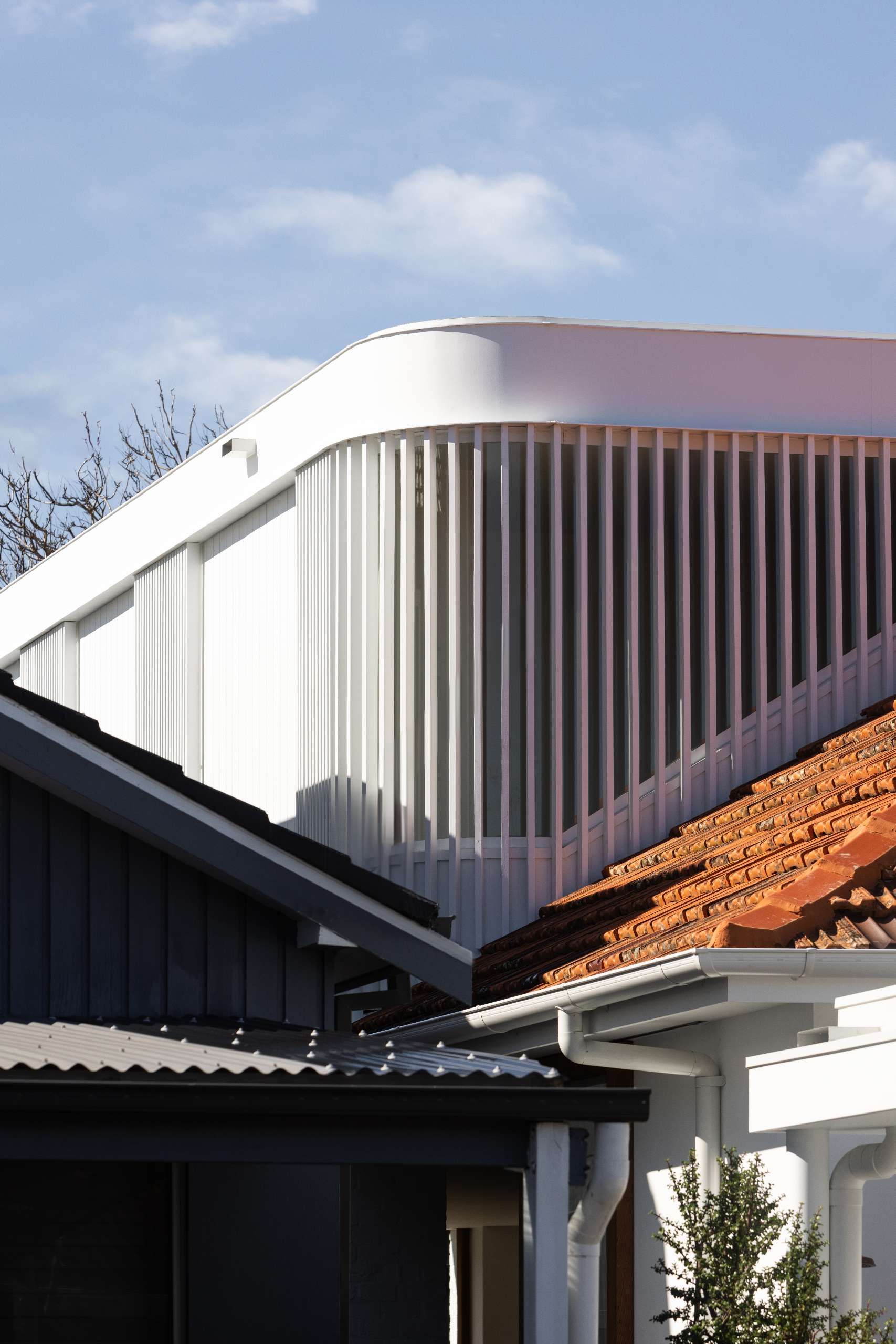 A curving white upper level with vertical battens rises above the traditional terracotta-roof, blending bold modernism with suburban heritage.