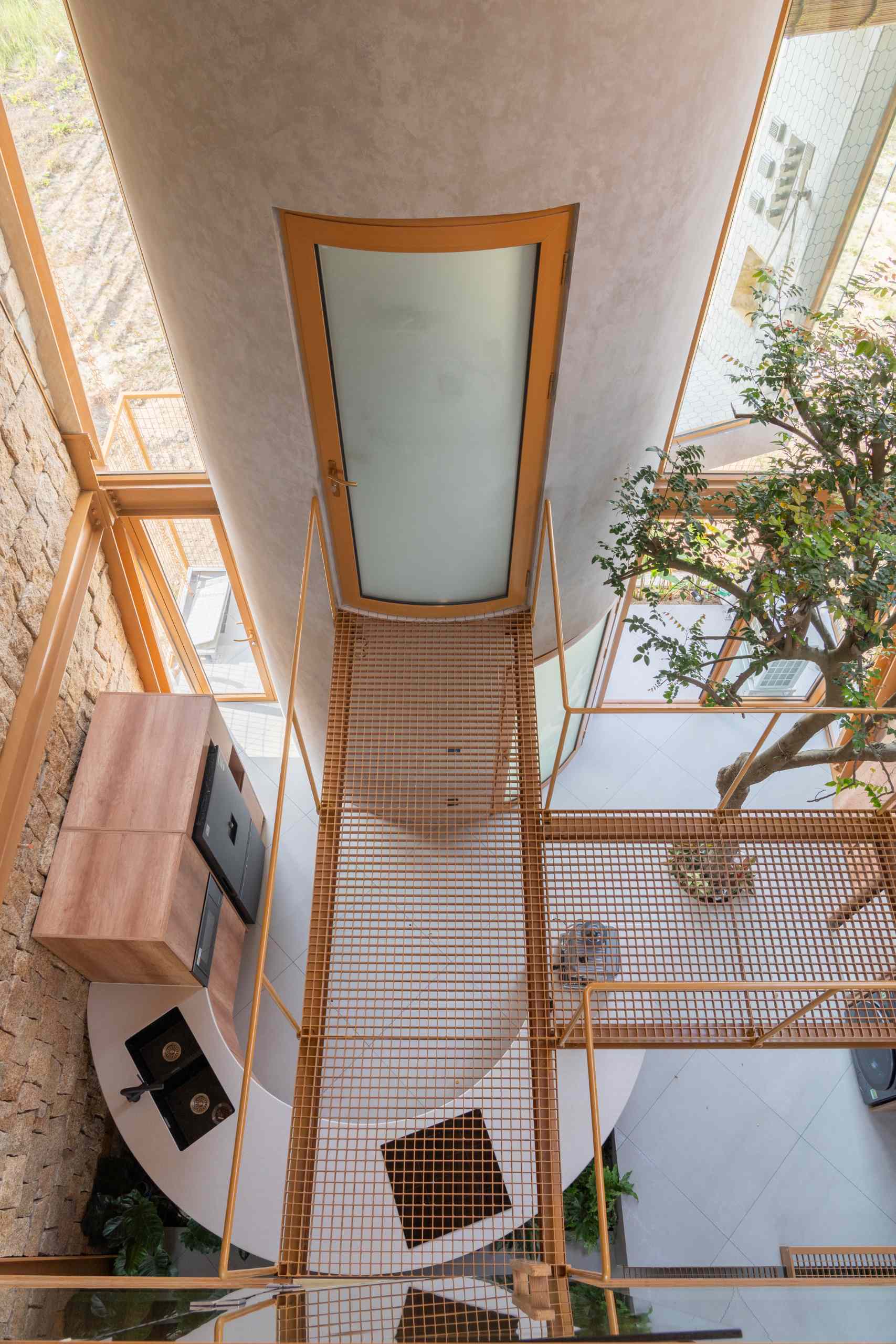 A metal walkway leads to a curved plastered bathroom volume, featuring a frosted glass door with a wood frame. Set between stone and glass walls, it blends raw textures with crisp, modern lines and a floating, elevated feel.
