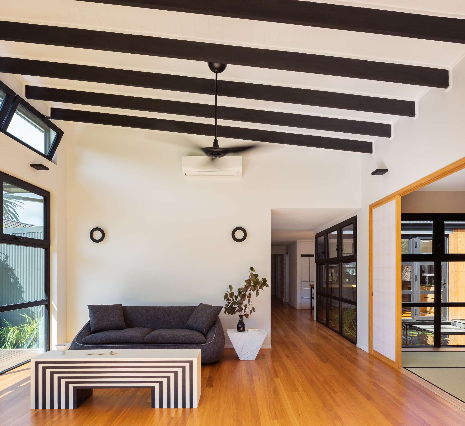 A bold yet tranquil living room blends Japanese minimalism with graphic modernism, exposed beams, a sculptural striped table, and black-framed windows offer strong contrasts softened by warm timber floors and a calming courtyard view.