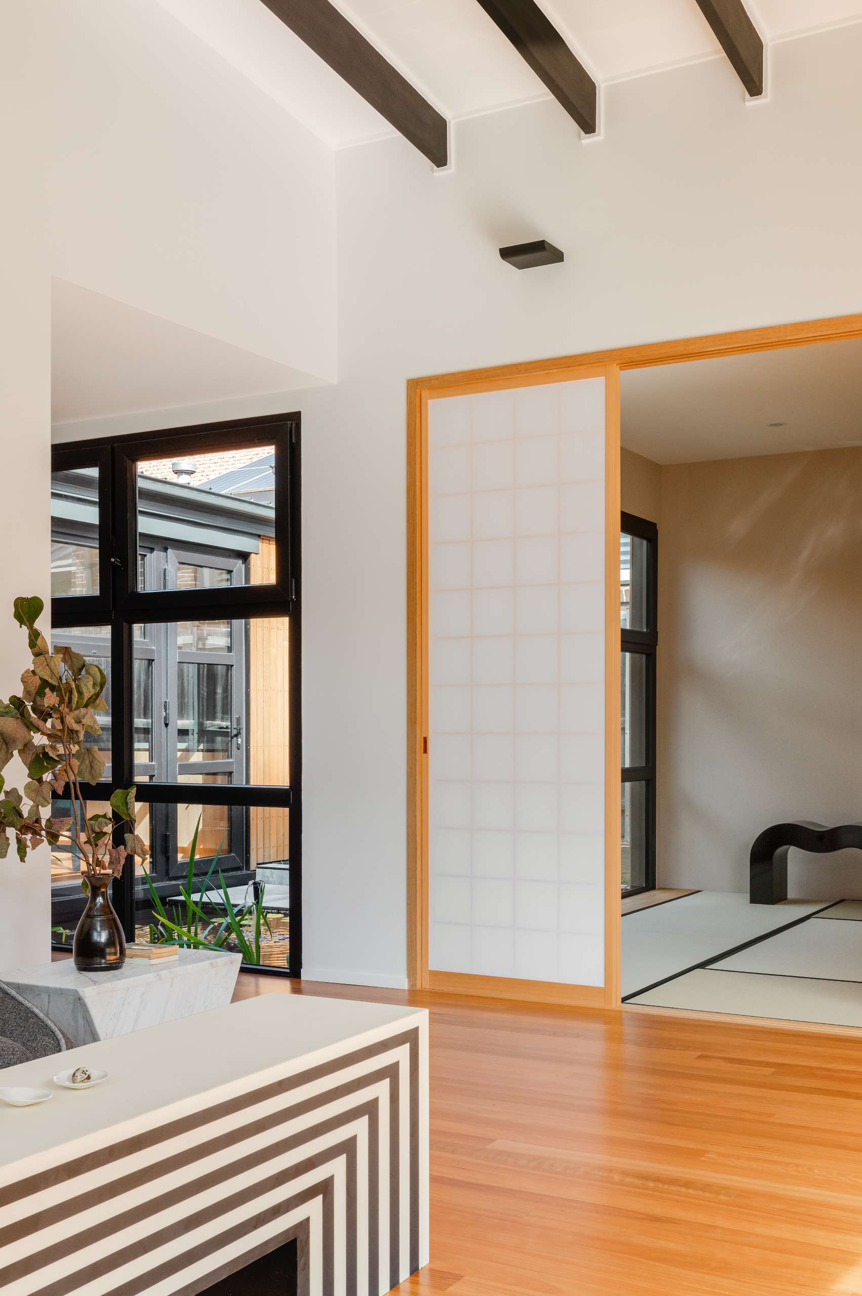 A bold yet tranquil living room blends Japanese minimalism with graphic modernism, exposed beams, a sculptural striped table, and black-framed windows offer strong contrasts softened by warm timber floors and a calming courtyard view.