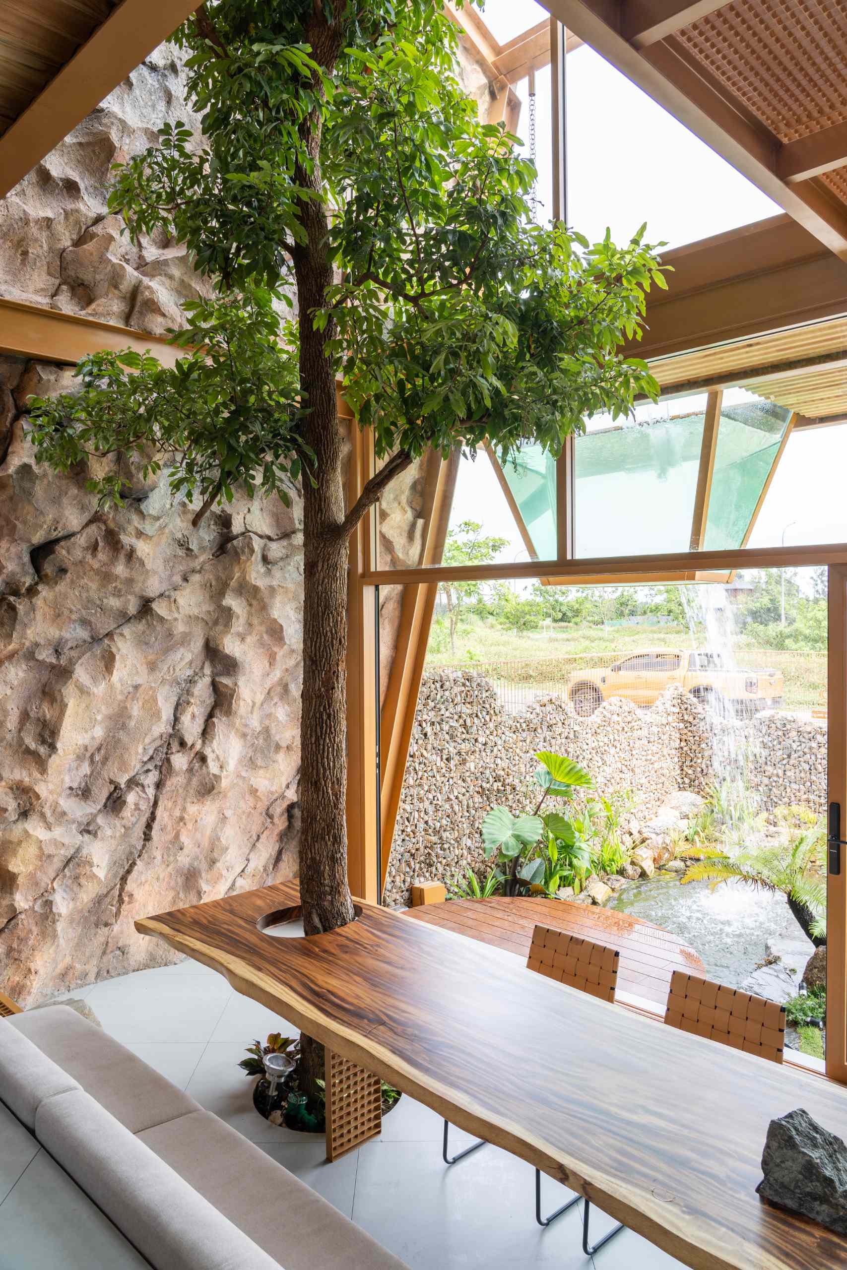 A full-grown tree rises through a custom wood slab table, blending nature with furniture in a playful, grounded way. The table’s rich grain and organic edge add warmth, while a stone base brings an earthy, solid touch to the space.