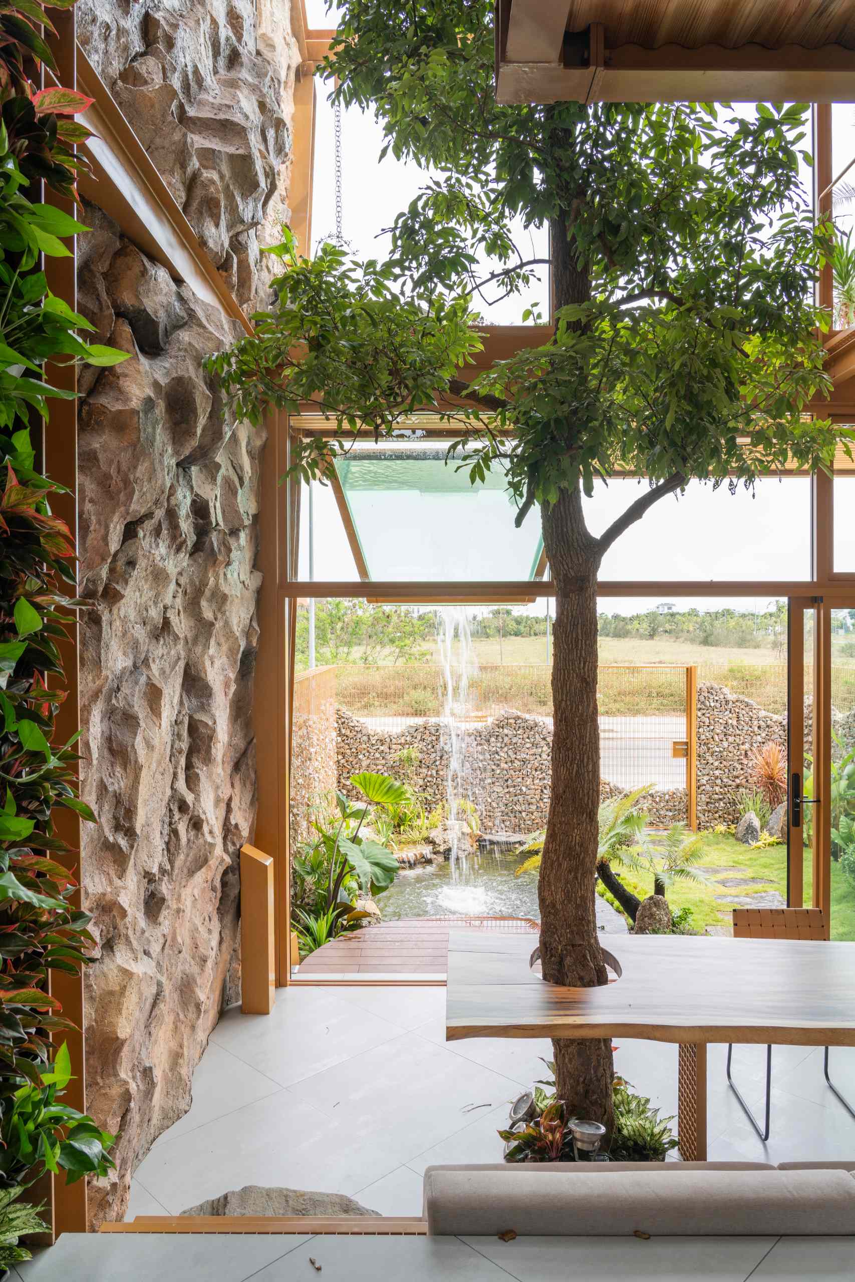A glass volume suspended above a pond creates a striking waterfall that cools the space and adds a calming sound. Surrounded by tropical plants, natural stone, and a textured gabion wall, the garden feels like a small, living ecosystem built into the heart of a modern home.