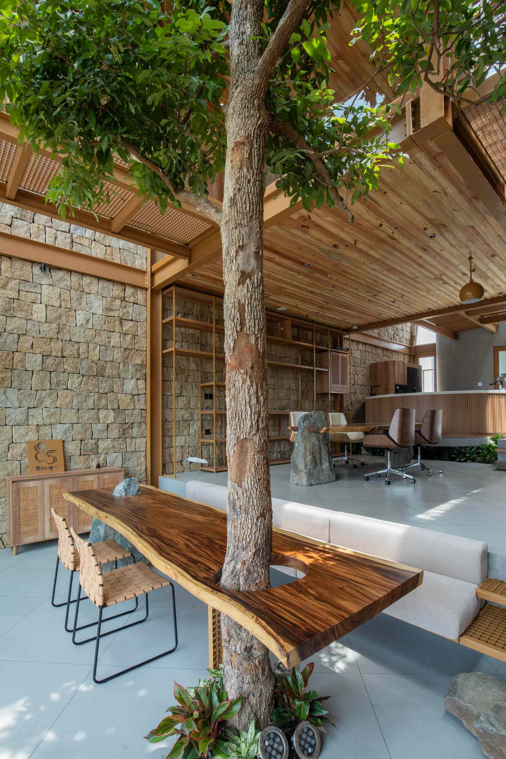 A full-grown tree rises through a custom wood slab table, blending nature with furniture in a playful, grounded way. The table’s rich grain and organic edge add warmth, while a stone base brings an earthy, solid touch to the space.