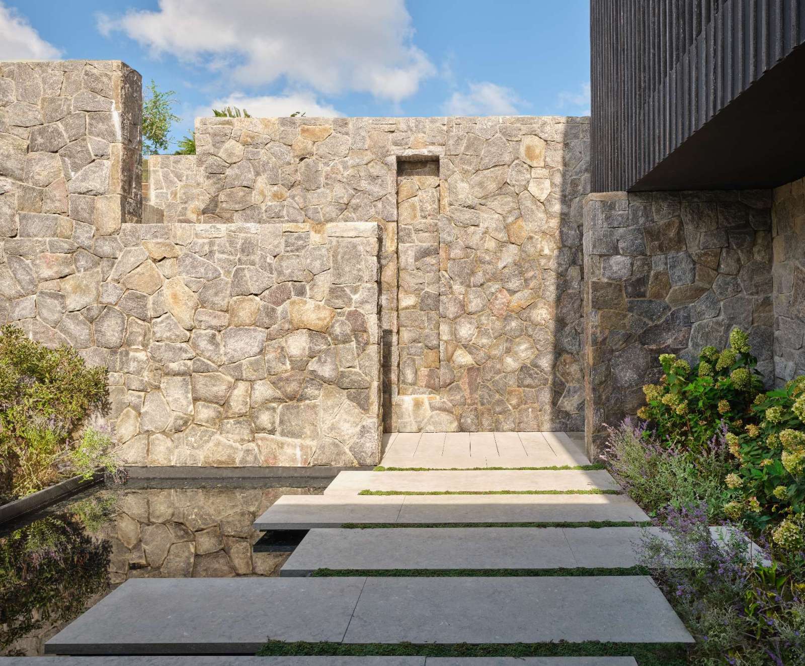 Stepping down towards the home, large pavers appear to float above a shallow reflective pool, framed by soft green moss that adds rhythm and contrast. Rugged stone walls anchor the space with texture and warmth, while the vertical wood slats filters sunlight and casts dynamic shadows.