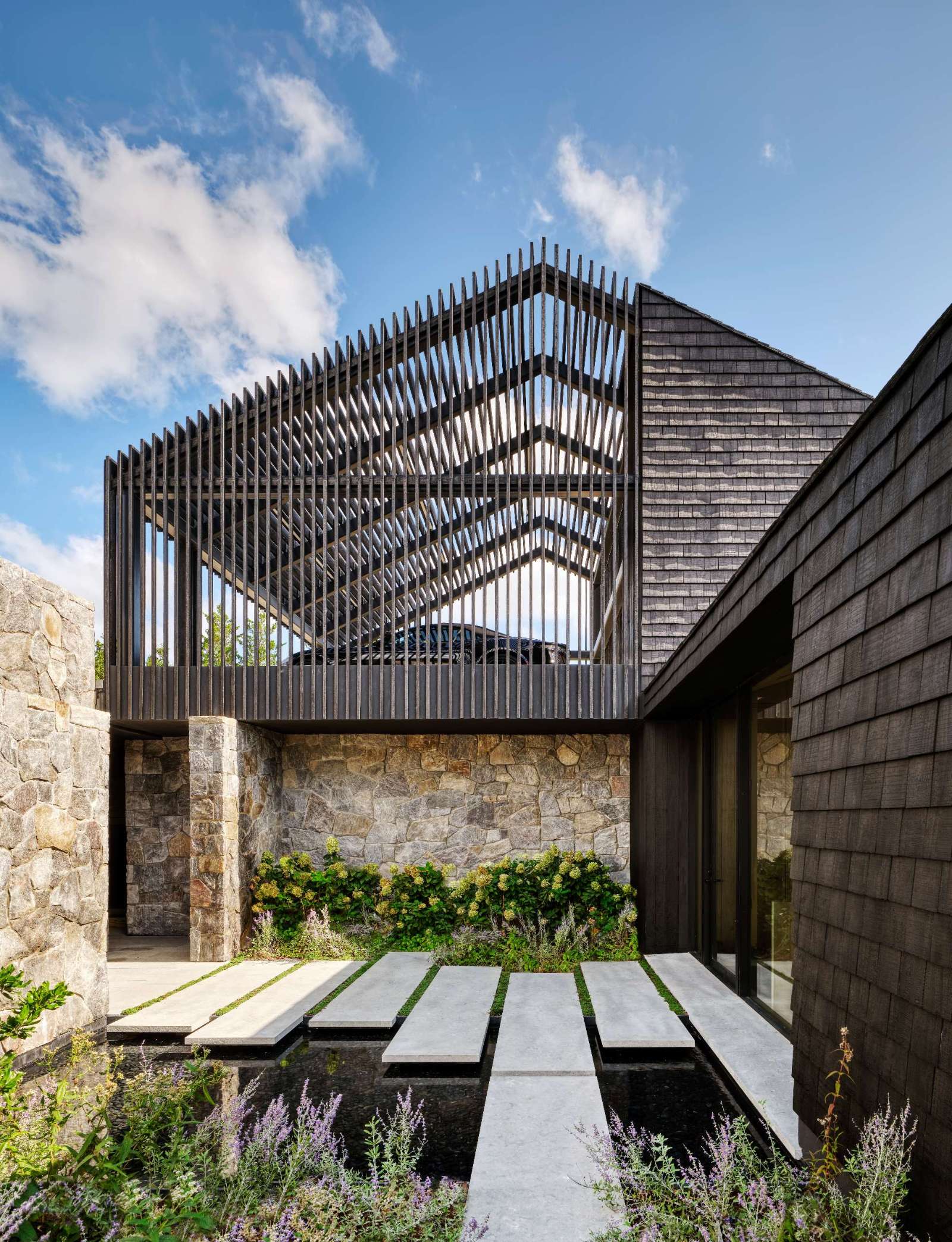 Stepping down towards the home, large pavers appear to float above a shallow reflective pool, framed by soft green moss that adds rhythm and contrast. Rugged stone walls anchor the space with texture and warmth, while the vertical wood slats filters sunlight and casts dynamic shadows.