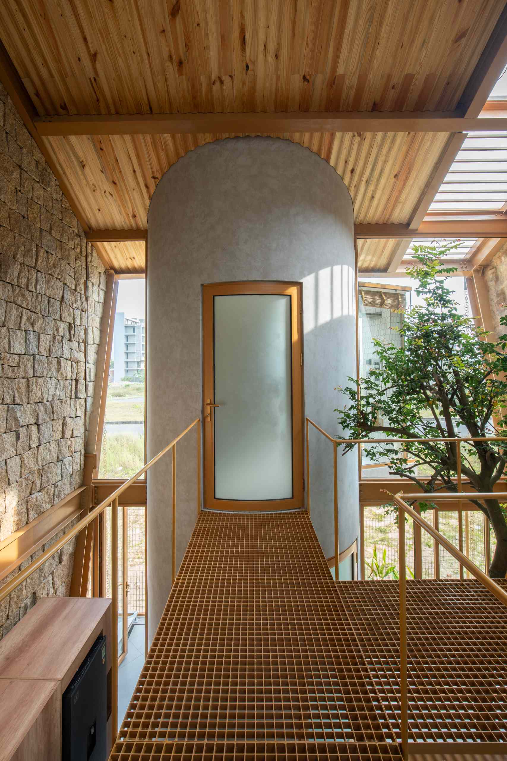A metal walkway leads to a curved plastered bathroom volume, featuring a frosted glass door with a wood frame. Set between stone and glass walls, it blends raw textures with crisp, modern lines and a floating, elevated feel.