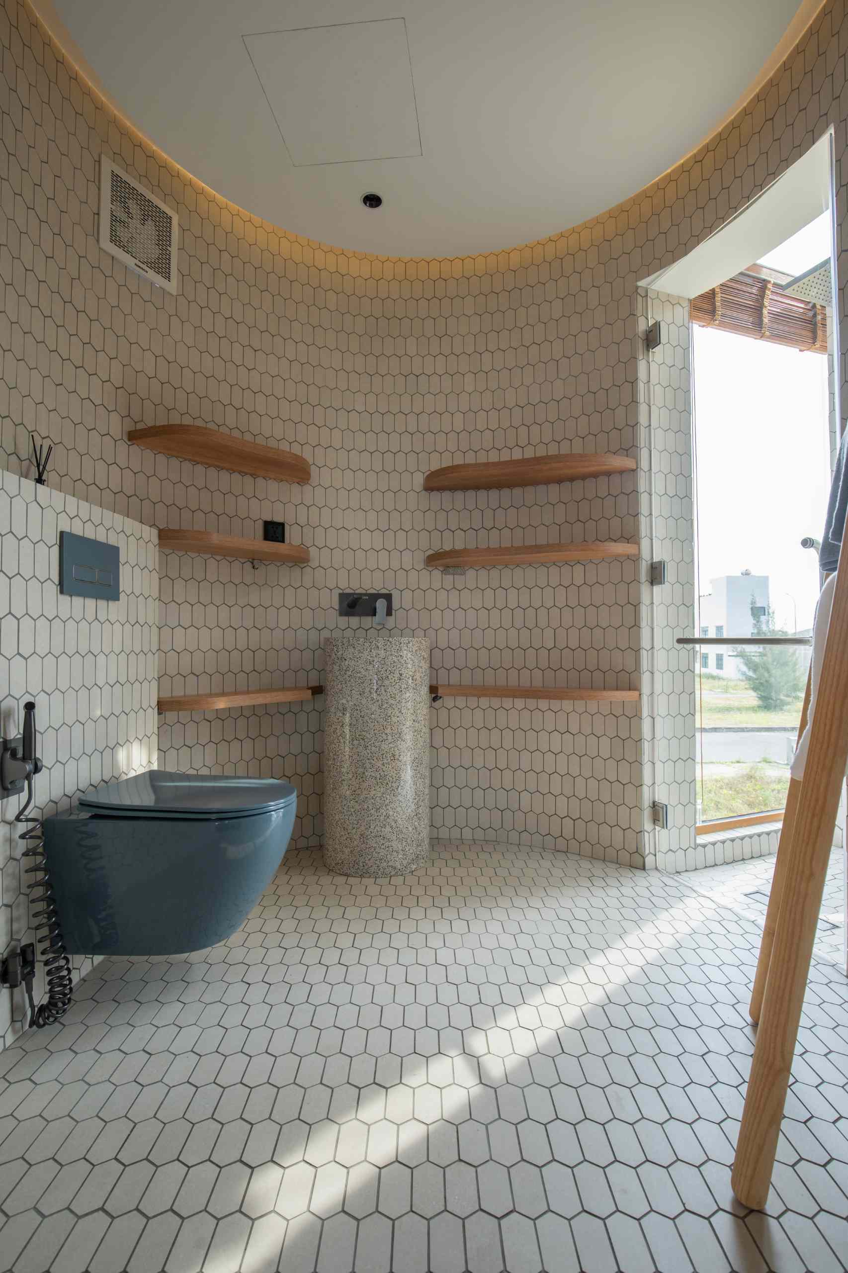 Cylindrical bathroom design featuring elongated hex tile walls and flooring, a sleek blue wall-mounted toilet, pedestal sink, floating wood shelves, and a glass-enclosed shower that fills the space with natural light.