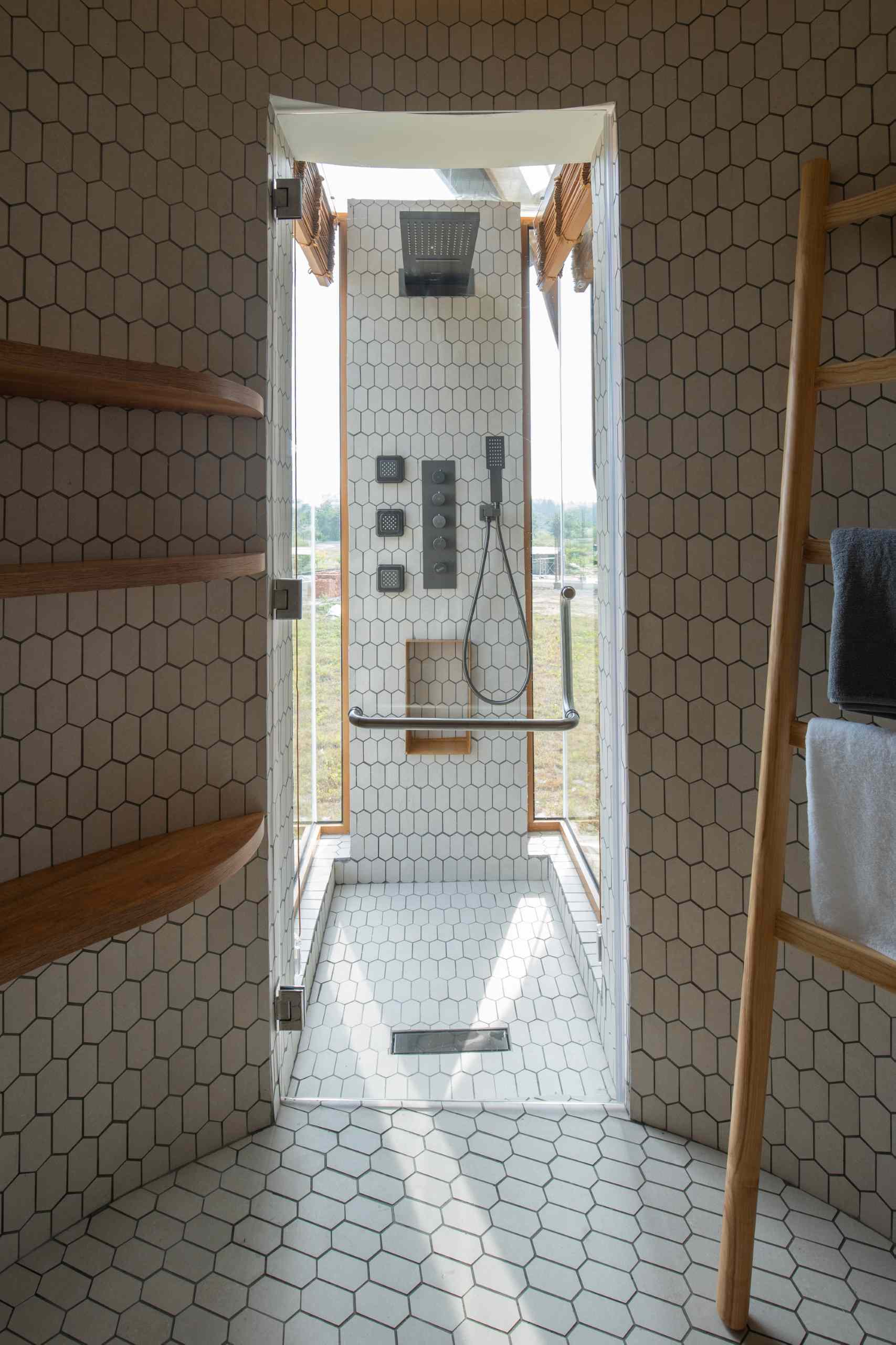 Cylindrical bathroom design featuring elongated hex tile walls and flooring, a sleek blue wall-mounted toilet, pedestal sink, floating wood shelves, and a glass-enclosed shower that fills the space with natural light.