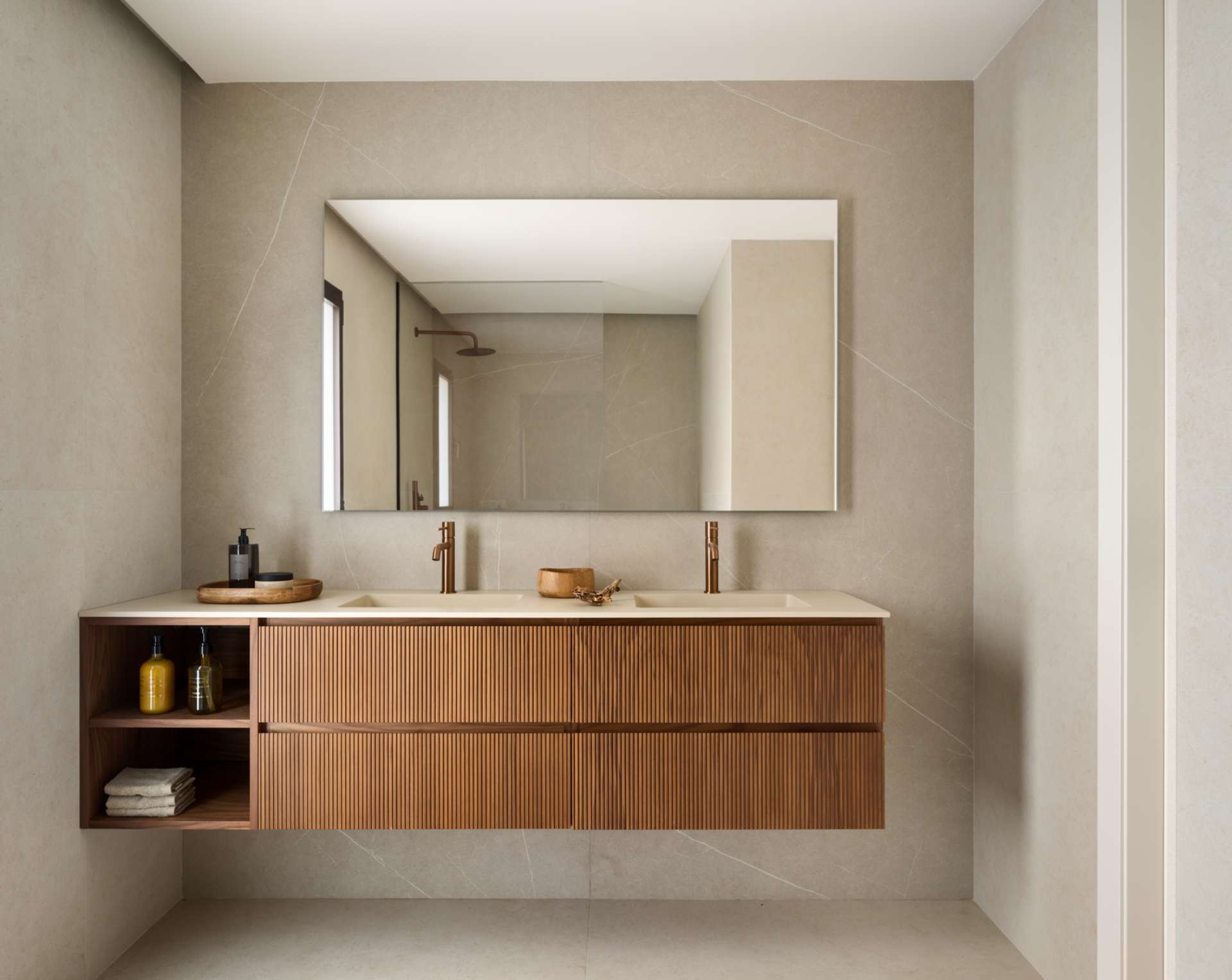 Clean lines and warm materials define the bathroom’s quiet elegance. A fluted timber vanity adds tactile interest beneath a wide mirror, while stone-look tiles and twin basins enhance the sense of space. Bronze fixtures introduce just a hint of glow, elevating the room without overpowering its simplicity.