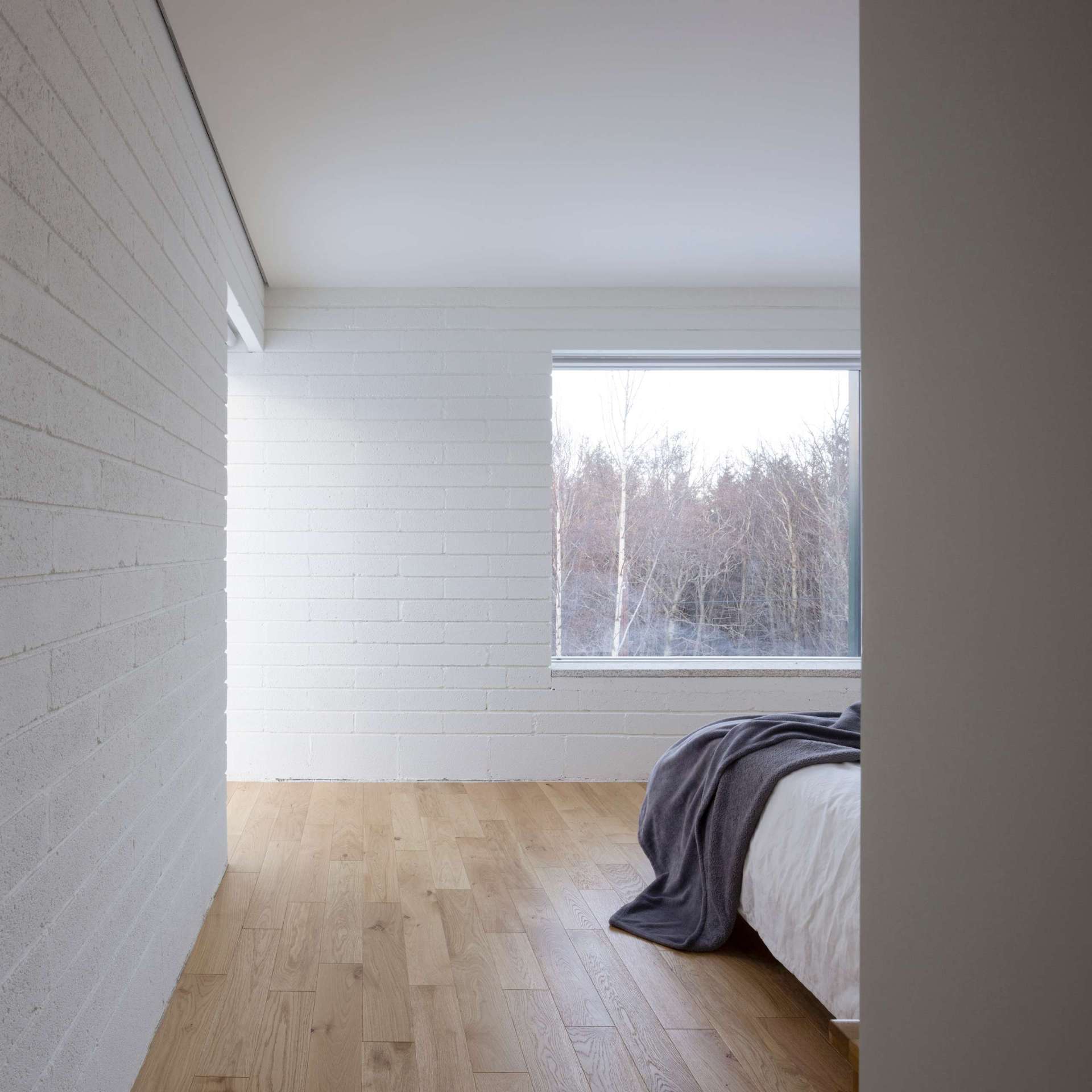A modern bedroom with wood flooring, a large window, and white-painted brick.