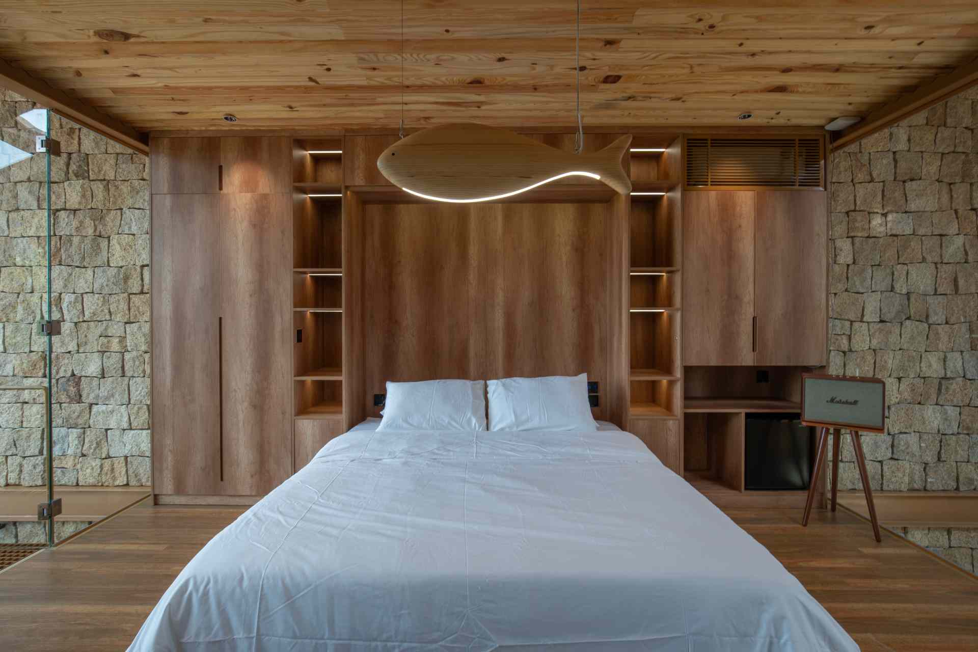 A light-filled home office enclosed in glass with views of a lush green wall. The wood-paneled wall behind the desk hides a fold-down bed, making it easy to switch from work mode to rest.