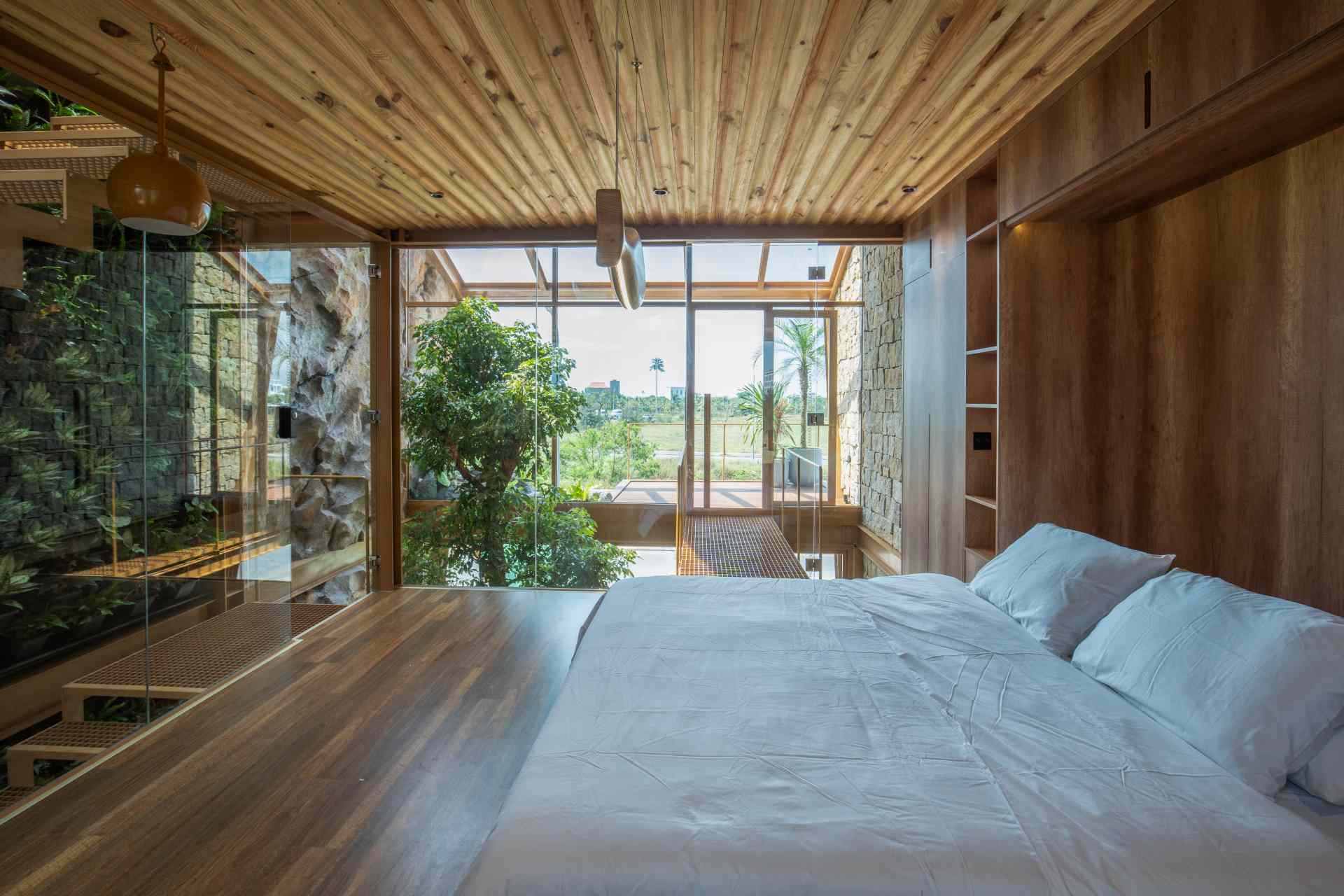 A light-filled home office enclosed in glass with views of a lush green wall. The wood-paneled wall behind the desk hides a fold-down bed, making it easy to switch from work mode to rest.