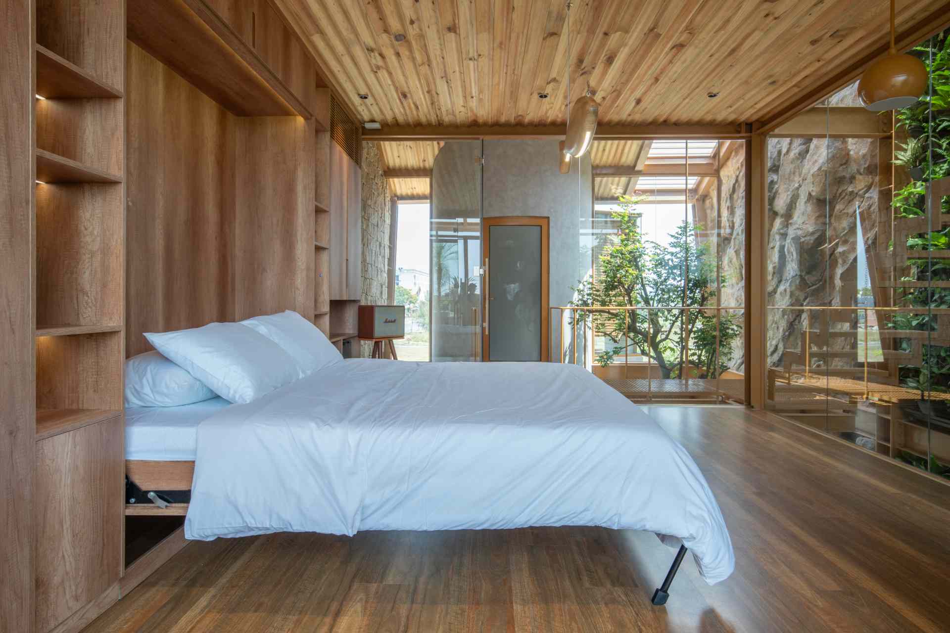 A light-filled home office enclosed in glass with views of a lush green wall. The wood-paneled wall behind the desk hides a fold-down bed, making it easy to switch from work mode to rest.